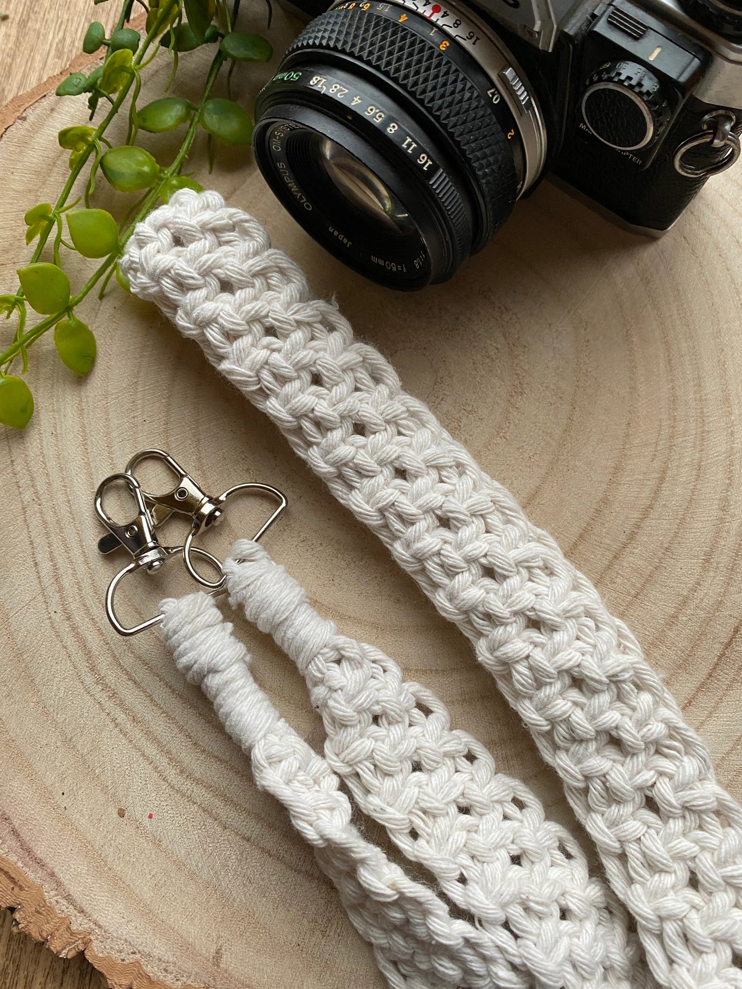 A black Olympus vintage camera lays on a round wooden surface, with a handmade macrame knotted camera strap next to it. The camera strap is made with natural coloured yarn and has metal clasps on each end.