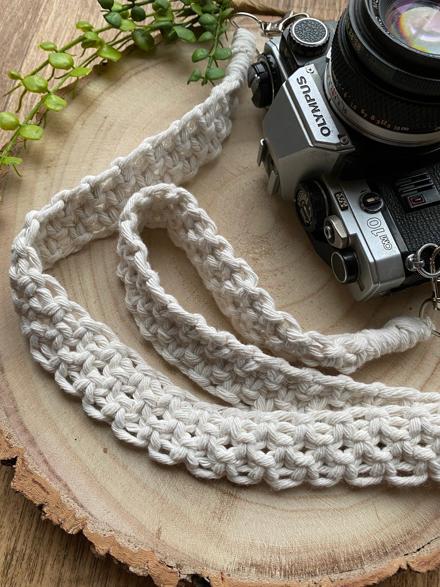 A black Olympus vintage camera lays on a round wooden surface, with a handmade macrame knotted camera strap attached to it with metal clasps. The camera strap is made with natural coloured yarn.