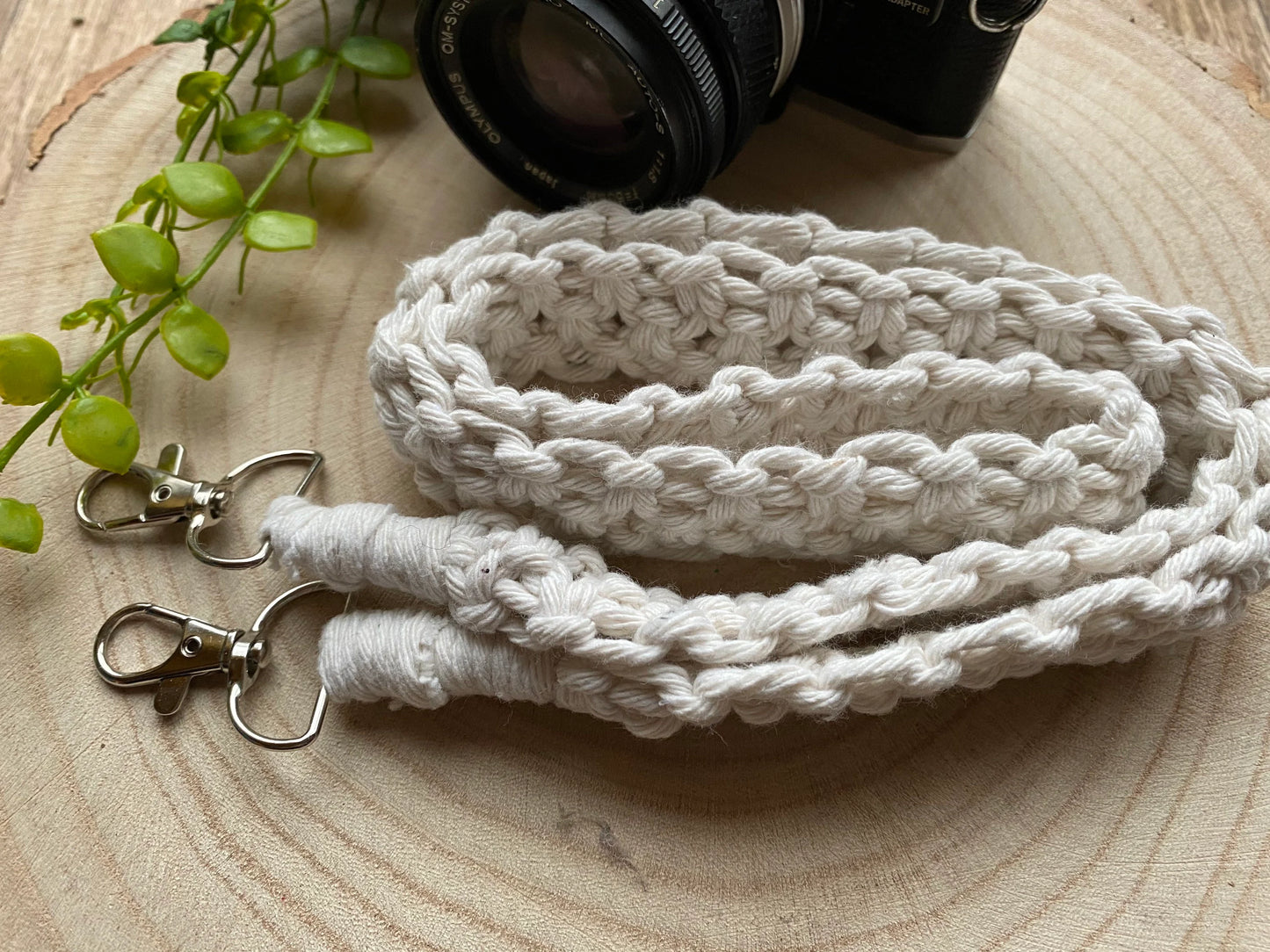 A handmade macrame knotted camera strap made with natural yarn, with metal clasps on each end, is coiled neatly on a round wooden surface, you can see the lens of an Olympus camera in the background.