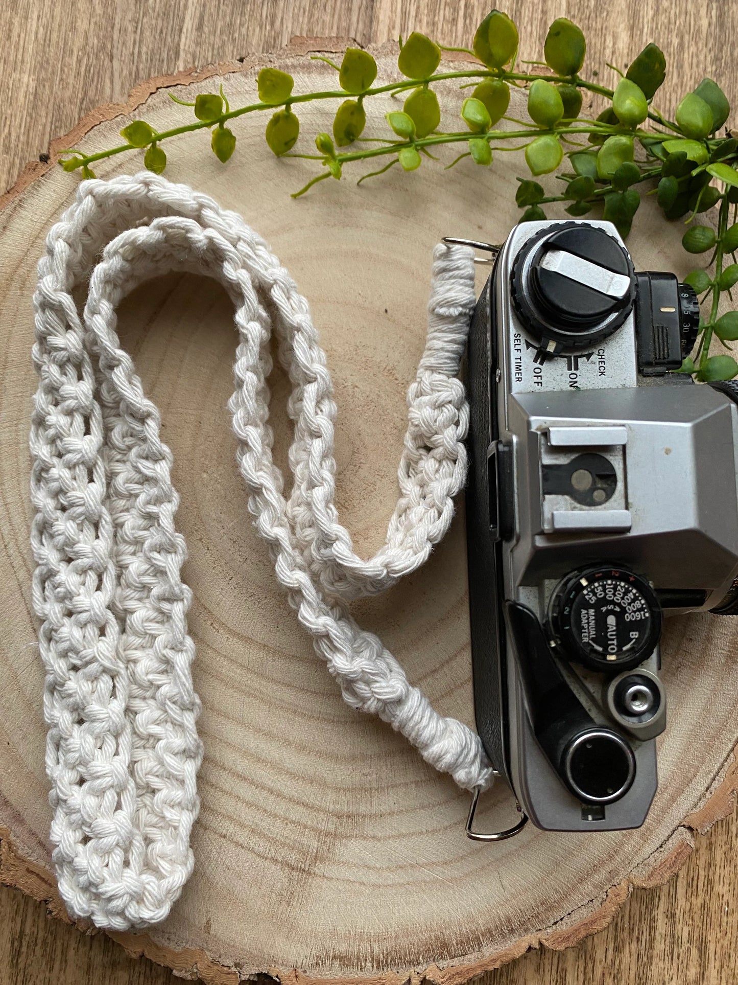 Top down view of a black Olympus vintage camera on a round wooden surface, with a handmade macrame knotted camera strap attached to it with metal clasps. The camera strap is made with natural yarn.