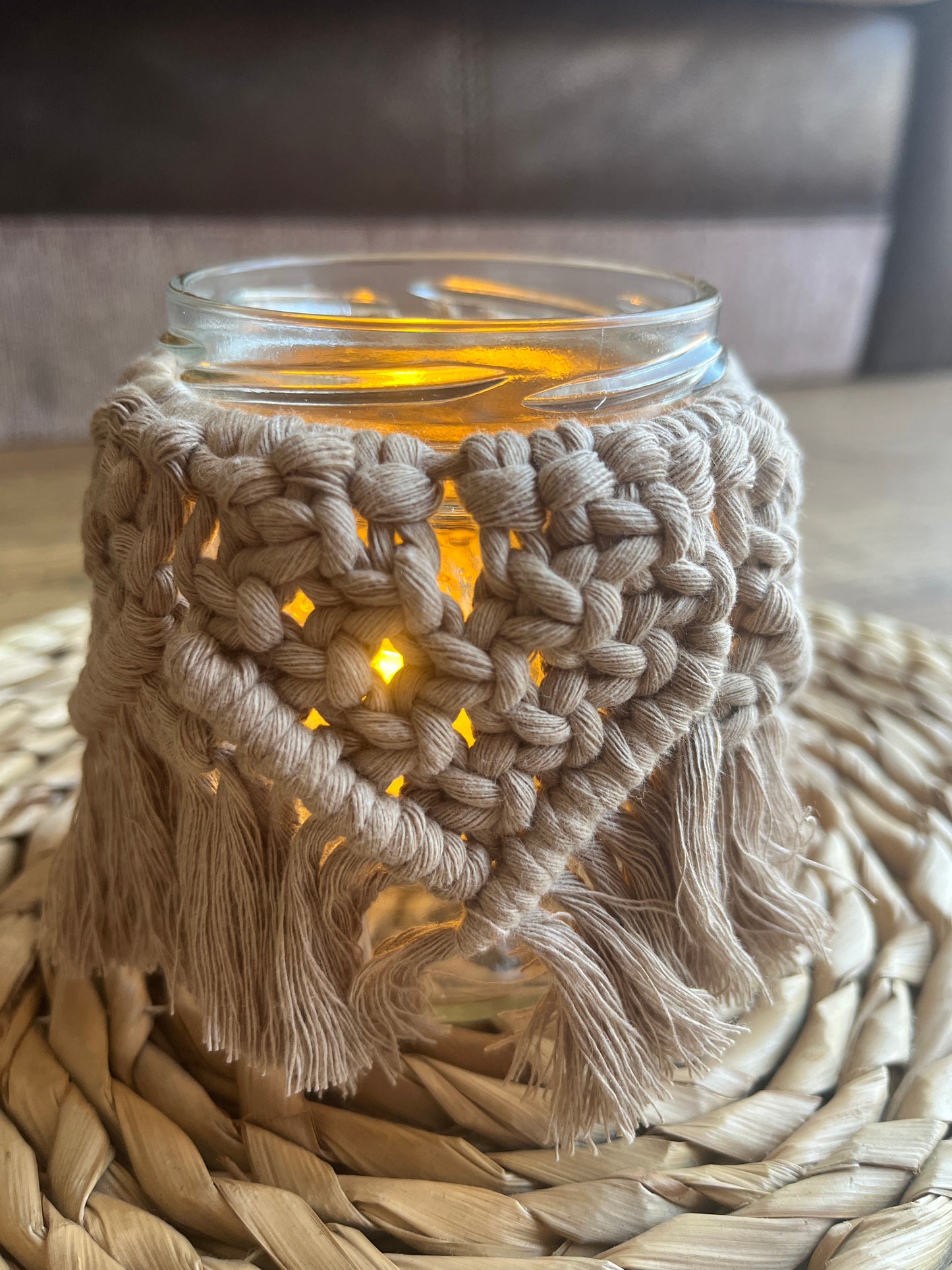 Close up of a glass jar wrapped with beige cotton yarn with a decorative knotted pattern. There is a soft glow from a candle coming from inside the jar.