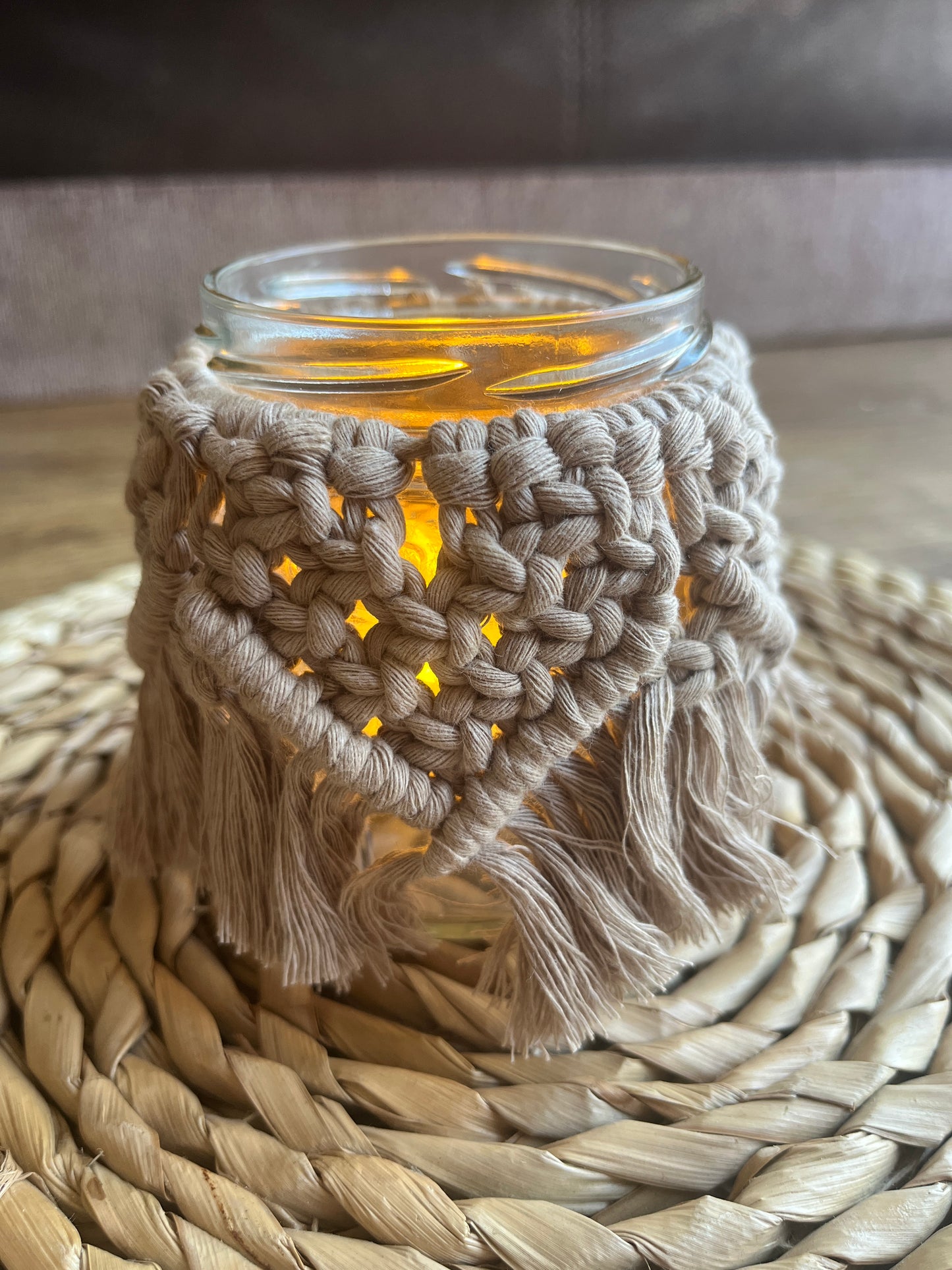 Close up of a glass jar wrapped with beige cotton yarn with a decorative knotted pattern placed on a wicker mat. There is a soft glow from a candle coming from inside the jar.