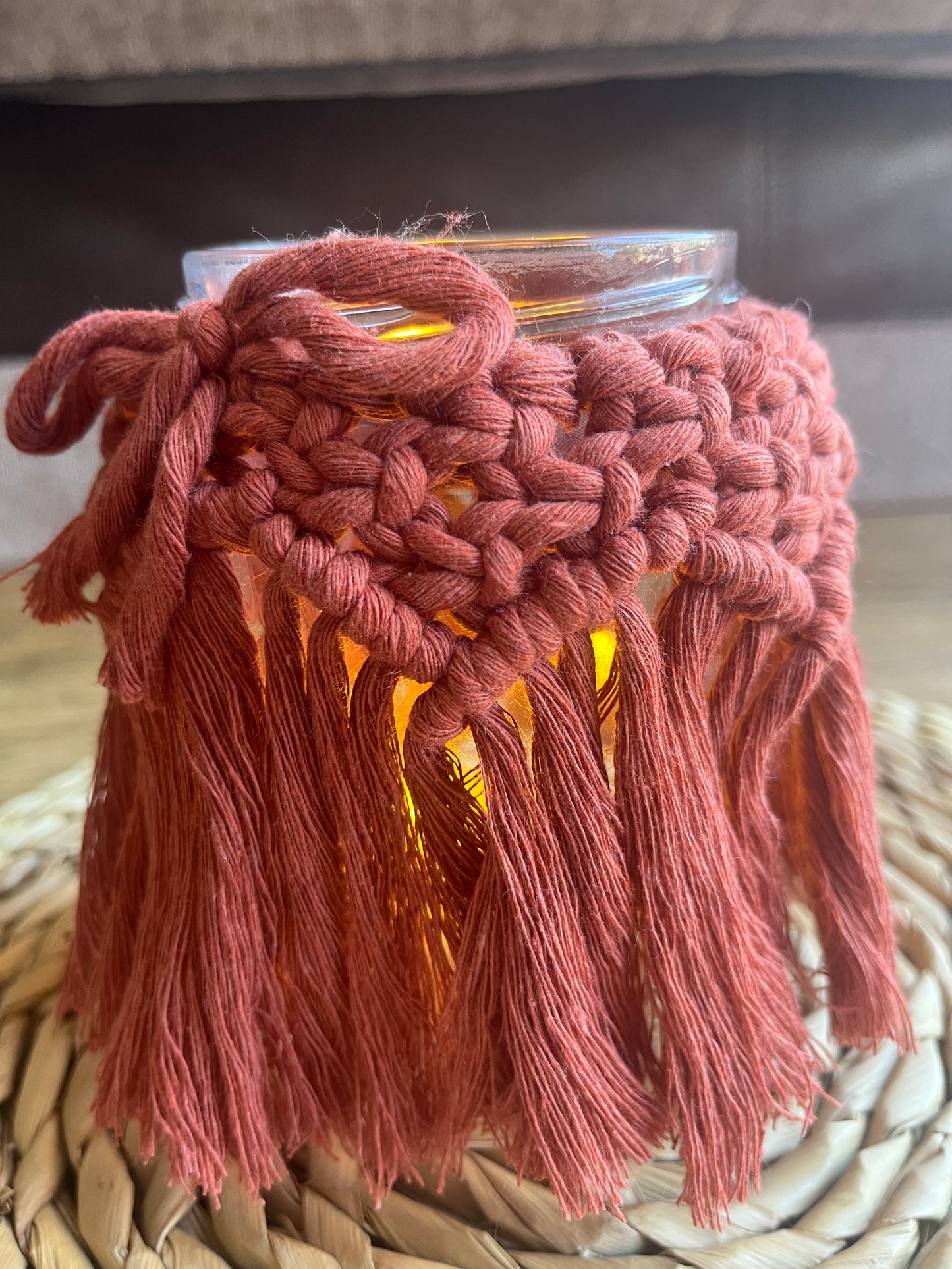 Close up of a glass jar wrapped with terracotta cotton yarn with a decorative knotted pattern and finished with a bow. There is a soft glow from a candle coming from inside the jar.