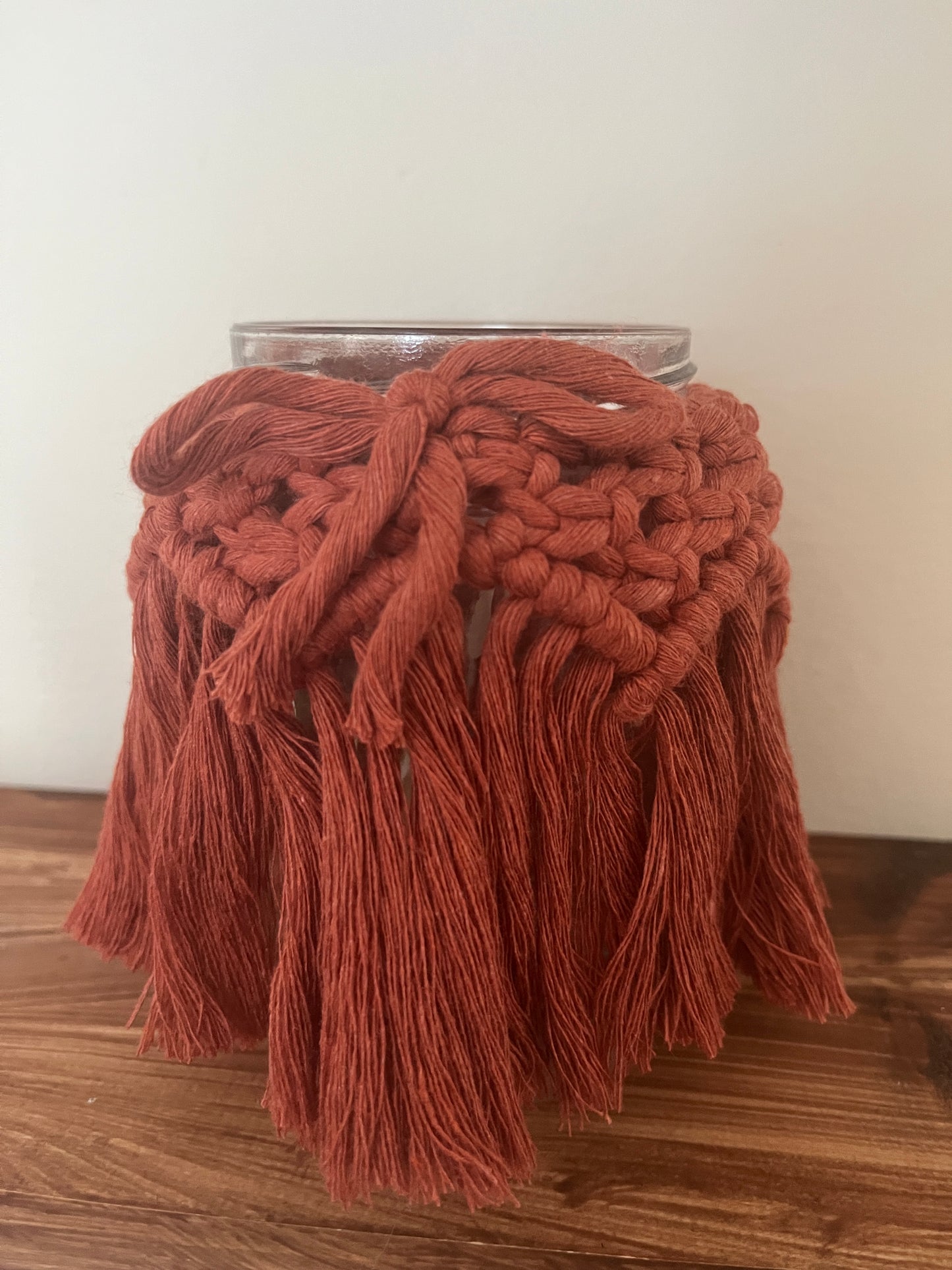 Close up of a glass jar wrapped with terracotta cotton yarn with a decorative knotted pattern and finished with a bow sitting on a wooden shelf.