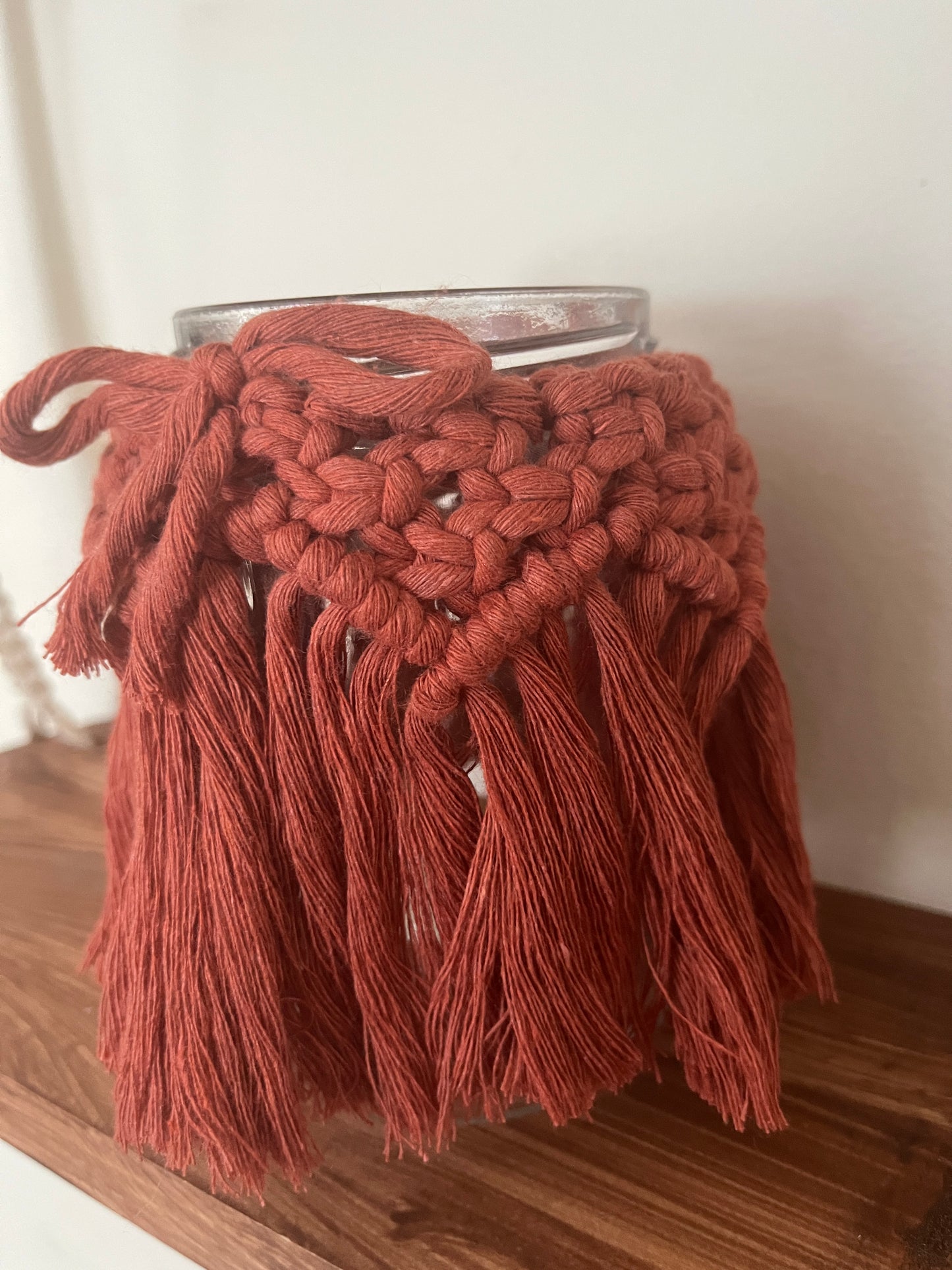 Close up of a glass jar wrapped with terracotta cotton yarn with a decorative knotted pattern and finished with a bow on a wooden shelf.