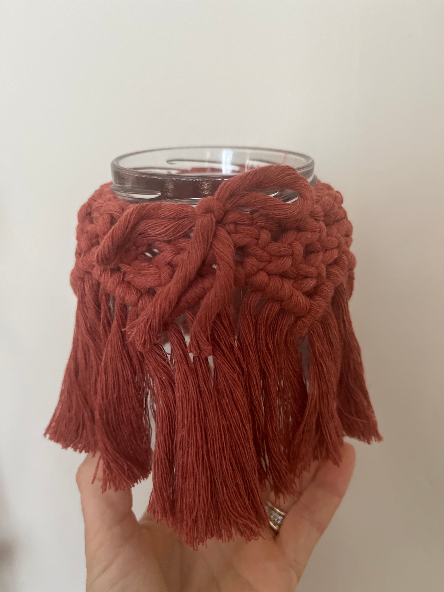 Hand holding a glass jar wrapped with terracotta cotton yarn with a decorative knotted pattern and finished with a bow, against a plain background.