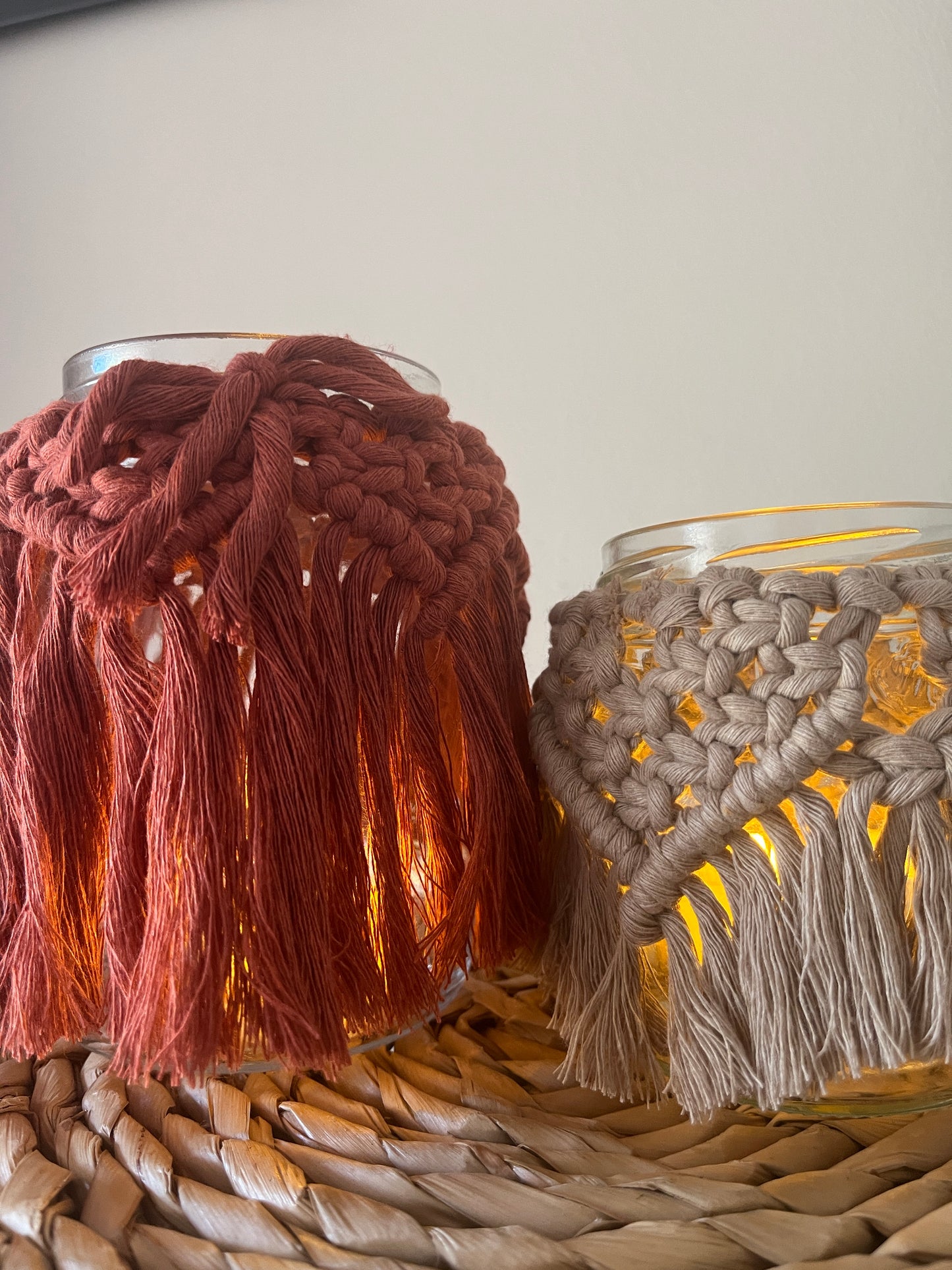 Two glass jars wrapped with cotton yarn with decorative knotted patterns placed close to one another on a wicker mat, in front of a plain background. There is a soft glow from a candle coming from inside the jars.