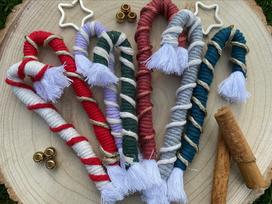 7 macrame candy cane Christmas decorations in a variety of colours, lay next to each other on a round wooden surface, surrounded by small beads and cinnamon sticks.
