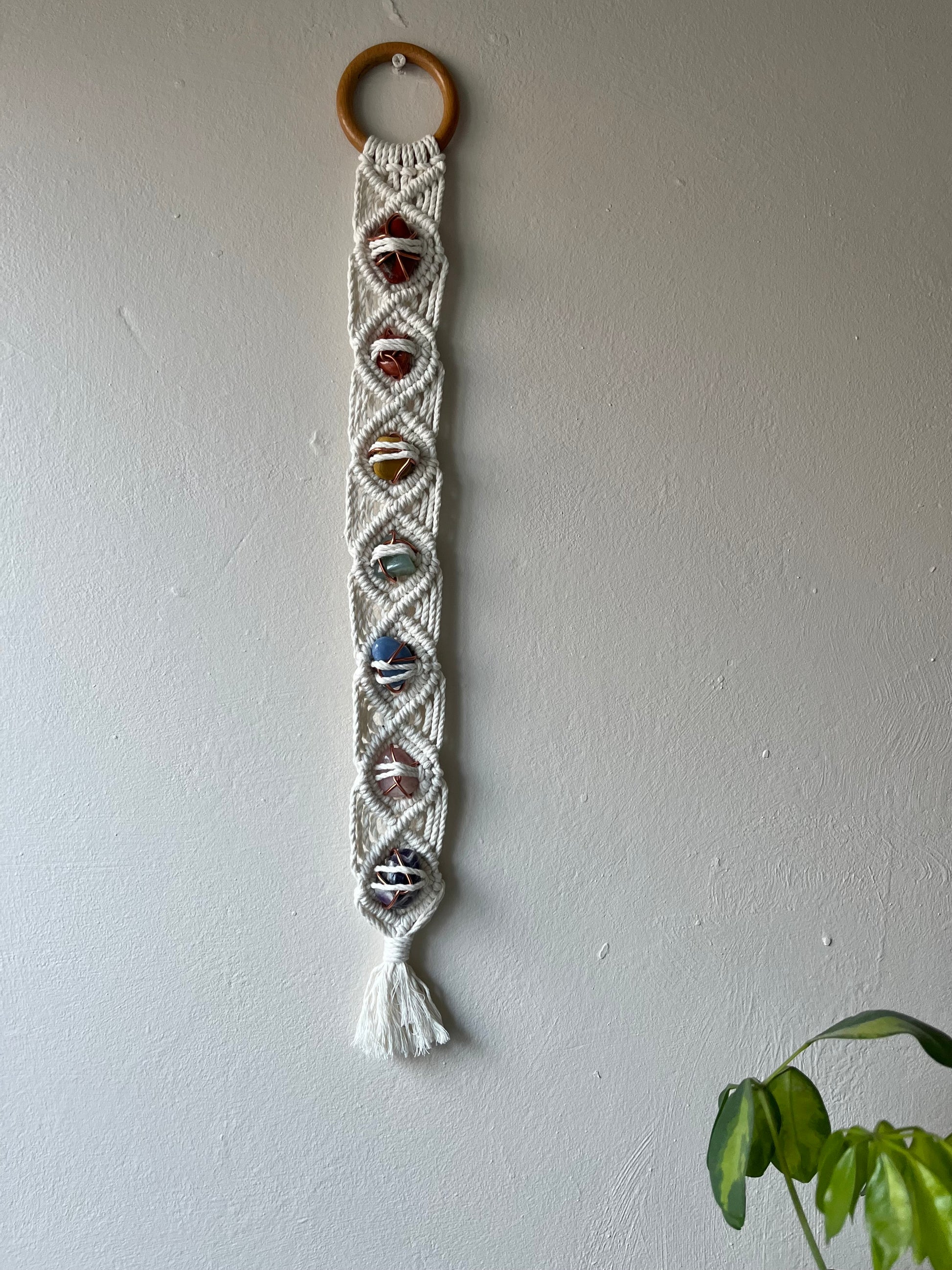 Long macrame wall hanging, featuring 7 gemstones to represent the chakras, held together with copper wire, and wrapped in natural recycled cotton yarn using a diamond knot pattern. finished with a repurposed wooden hanging ring. Displayed hanging on a white painted wall.