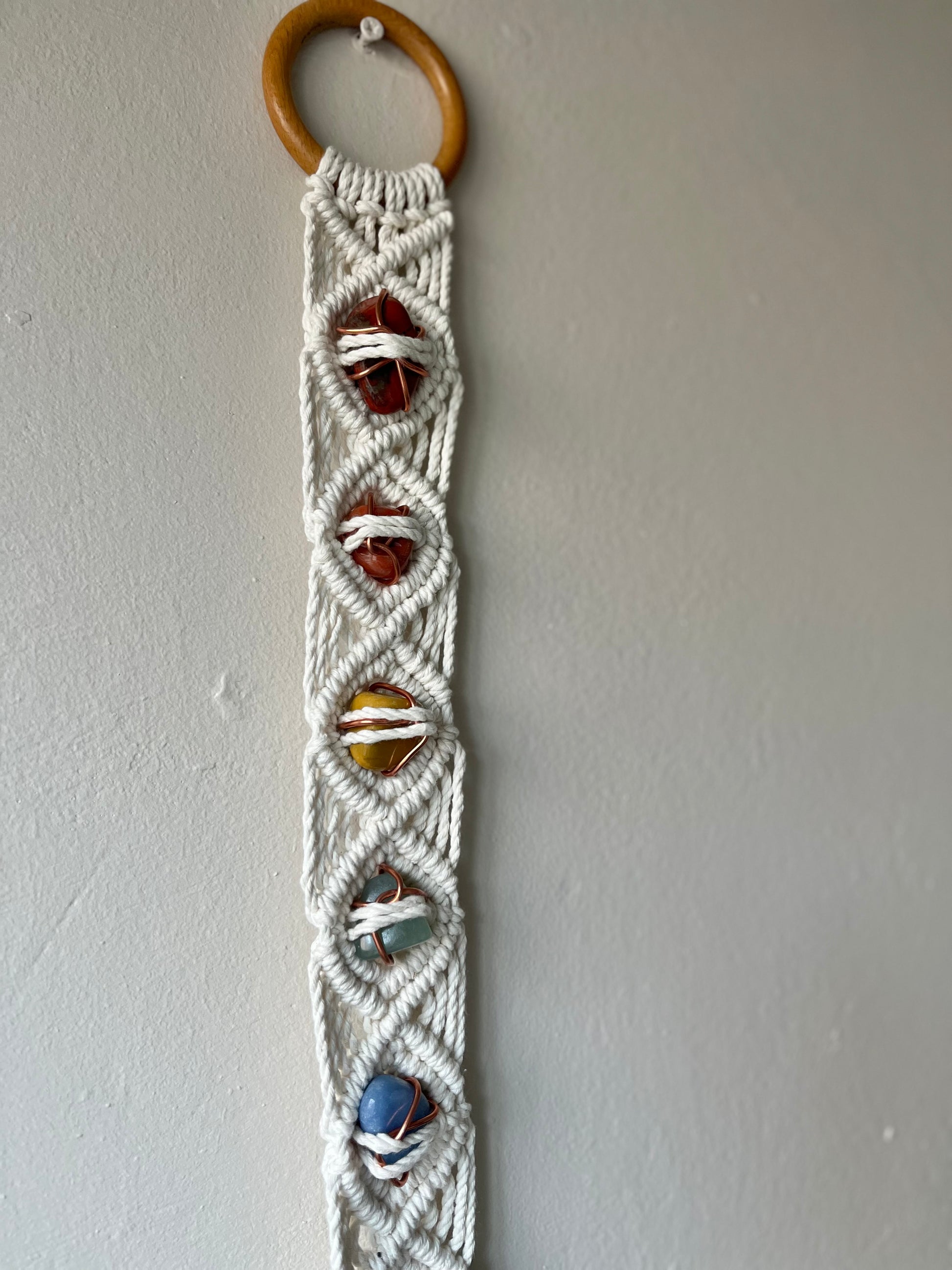 Long macrame wall hanging, featuring 7 gemstones to represent the chakras, held together with copper wire, and wrapped in natural recycled cotton yarn using a diamond knot pattern. finished with a repurposed wooden hanging ring. Displayed hanging on a white painted wall.