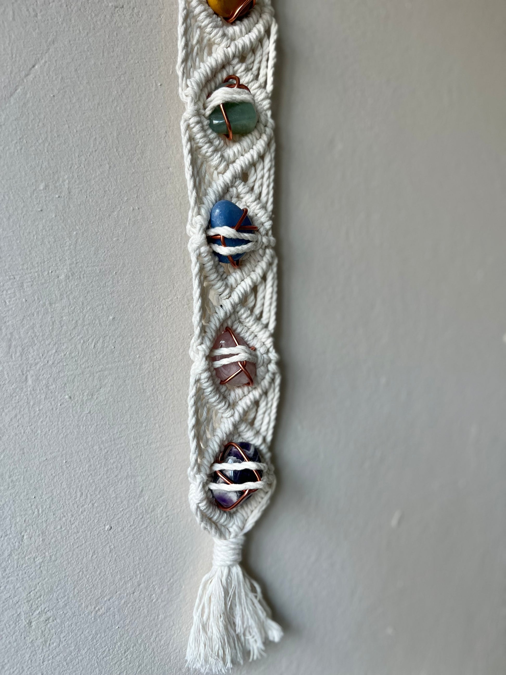 Long macrame wall hanging, featuring 7 gemstones to represent the chakras, held together with copper wire, and wrapped in natural recycled cotton yarn using a diamond knot pattern. finished with a repurposed wooden hanging ring. Displayed hanging on a white painted wall.
