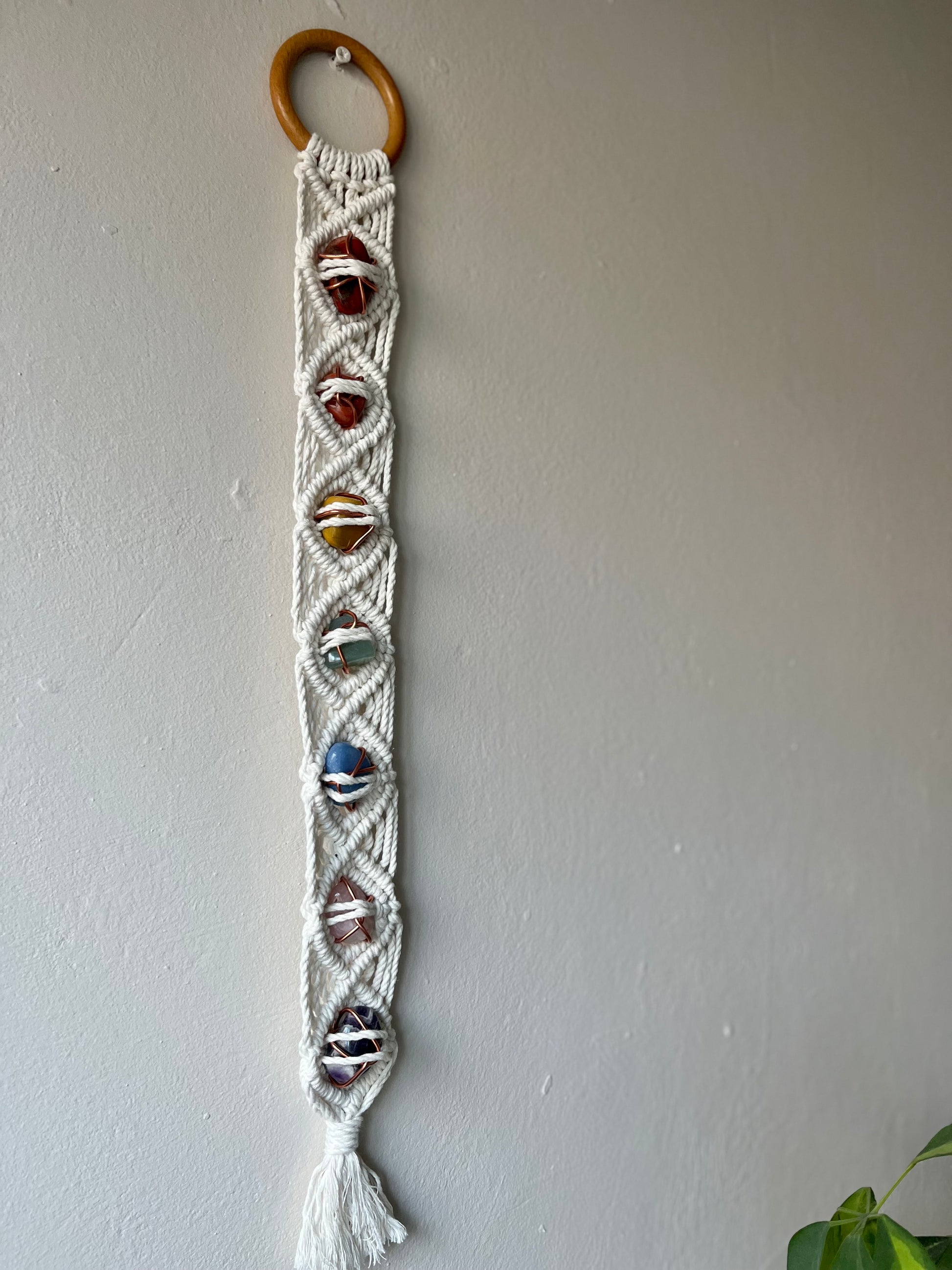 Long macrame wall hanging, featuring 7 gemstones to represent the chakras, held together with copper wire, and wrapped in natural recycled cotton yarn using a diamond knot pattern. finished with a repurposed wooden hanging ring. Displayed hanging on a white painted wall.