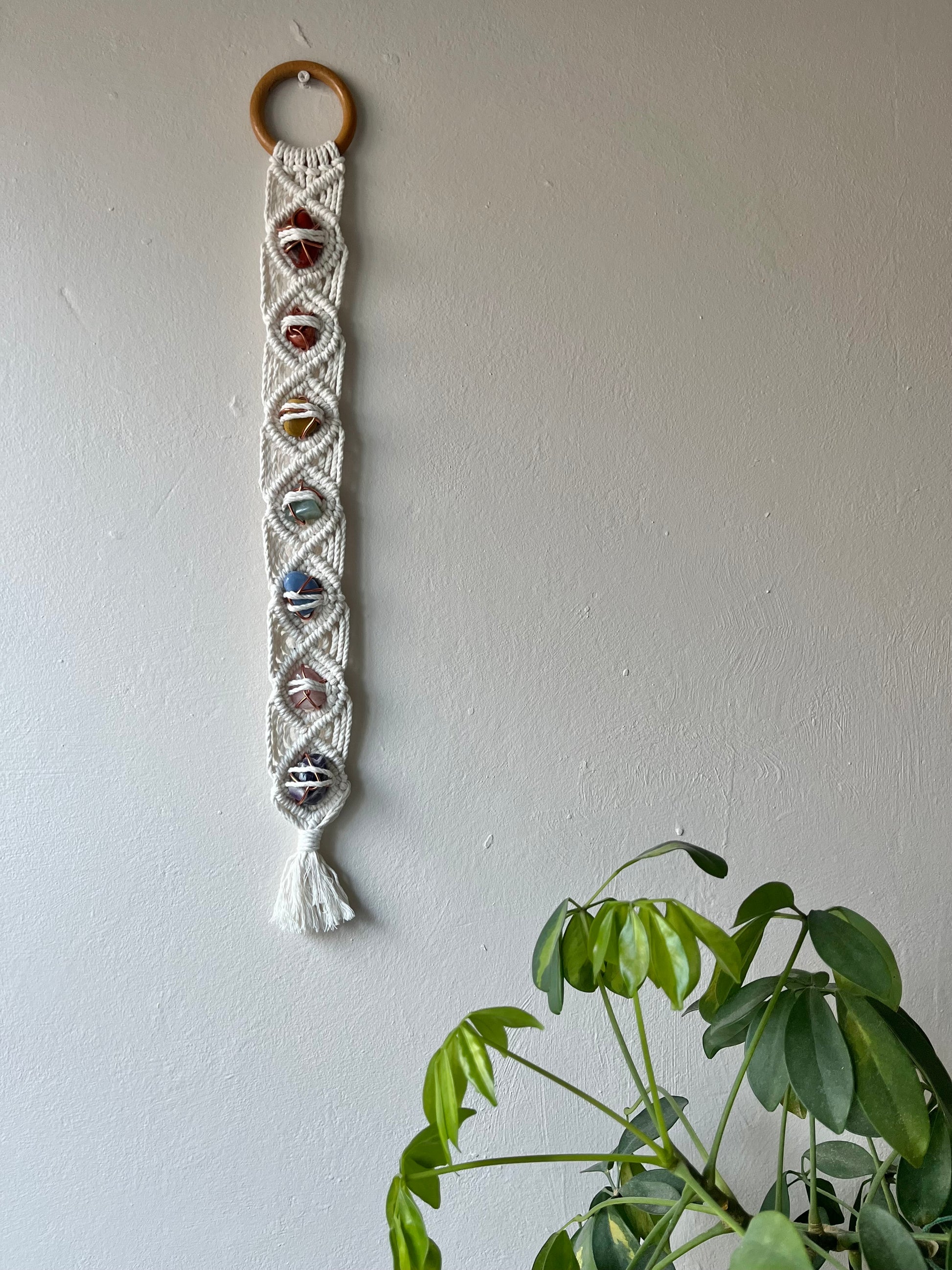 Long macrame wall hanging, featuring 7 gemstones to represent the chakras, held together with copper wire, and wrapped in natural recycled cotton yarn using a diamond knot pattern. finished with a repurposed wooden hanging ring. Displayed hanging on a white painted wall next to a large green plant.