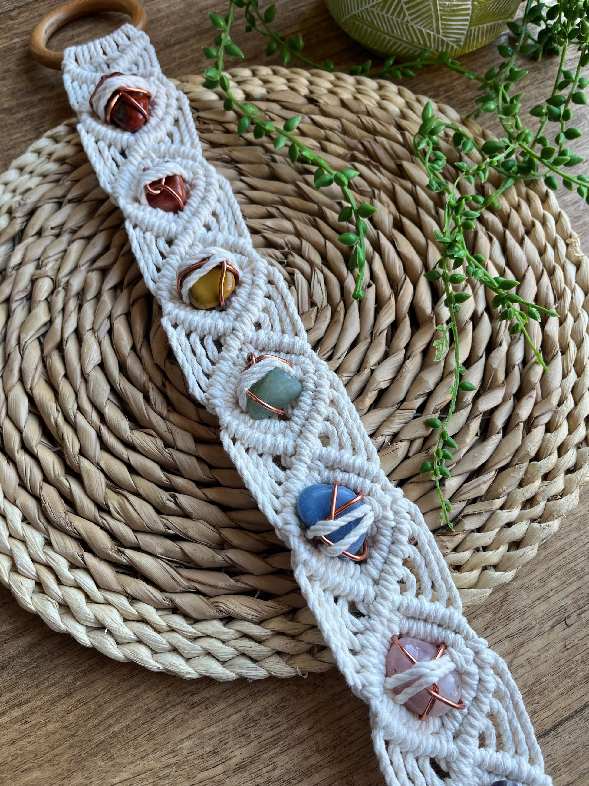 Long macrame wall hanging, featuring 7 gemstones to represent the chakras, held together with copper wire, and wrapped in natural recycled cotton yarn using a diamond knot pattern. finished with a repurposed wooden hanging ring. Displayed on a wicker mat with some green plant vines.