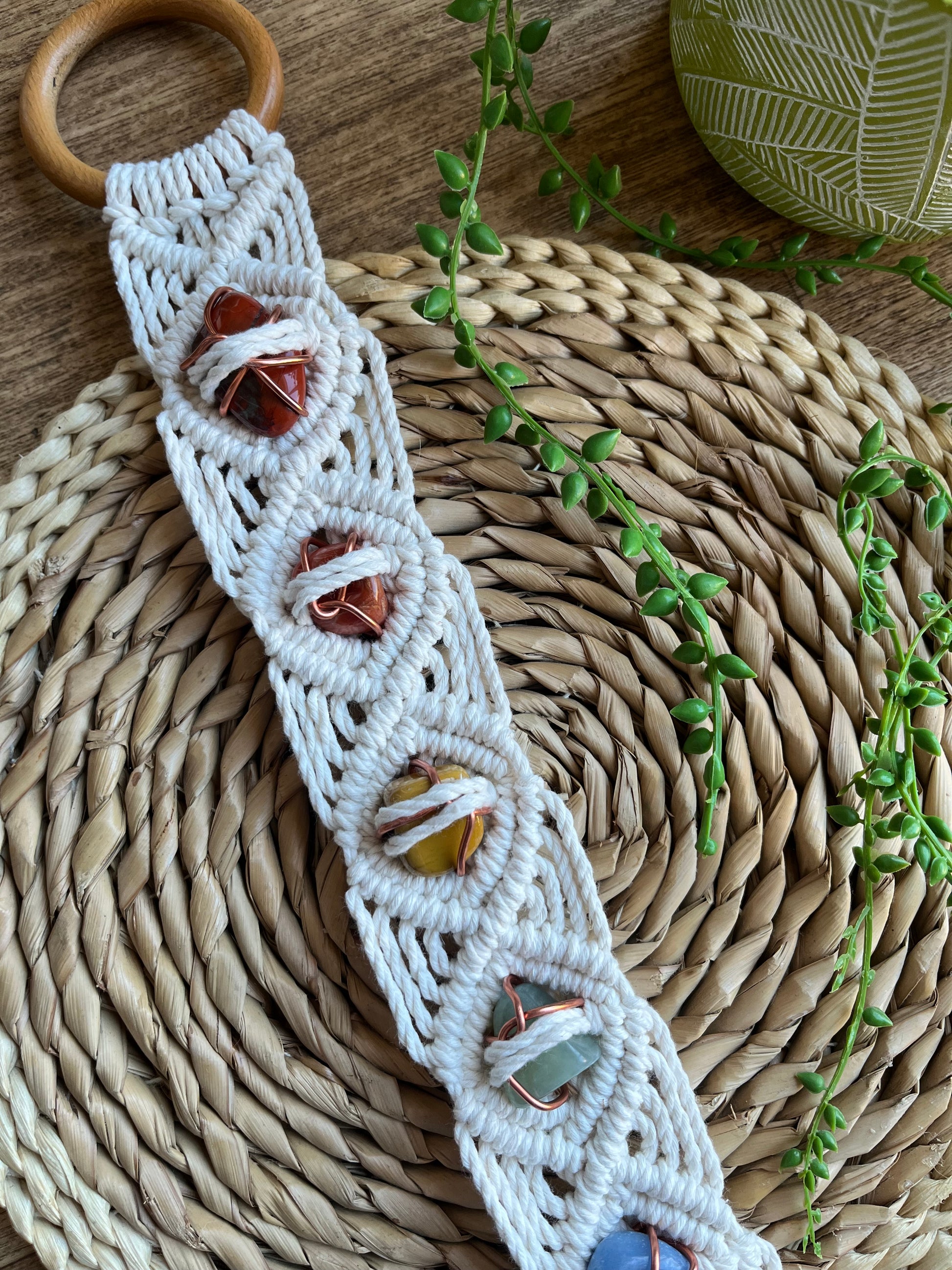 Long macrame wall hanging, featuring 7 gemstones to represent the chakras, held together with copper wire, and wrapped in natural recycled cotton yarn using a diamond knot pattern. finished with a repurposed wooden hanging ring. Displayed on a wicker mat with some green plant vines.