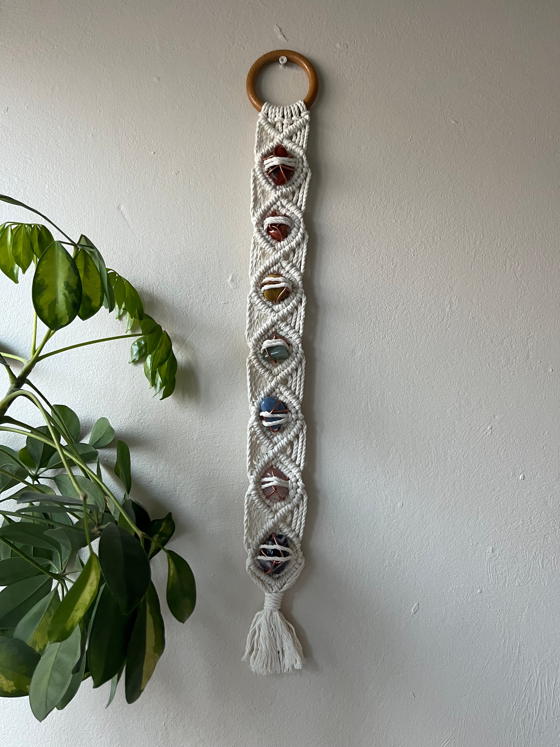 Long macrame wall hanging, featuring 7 gemstones to represent the chakras, held together with copper wire, and wrapped in natural recycled cotton yarn using a diamond knot pattern. finished with a repurposed wooden hanging ring. Displayed hanging on a white painted wall.