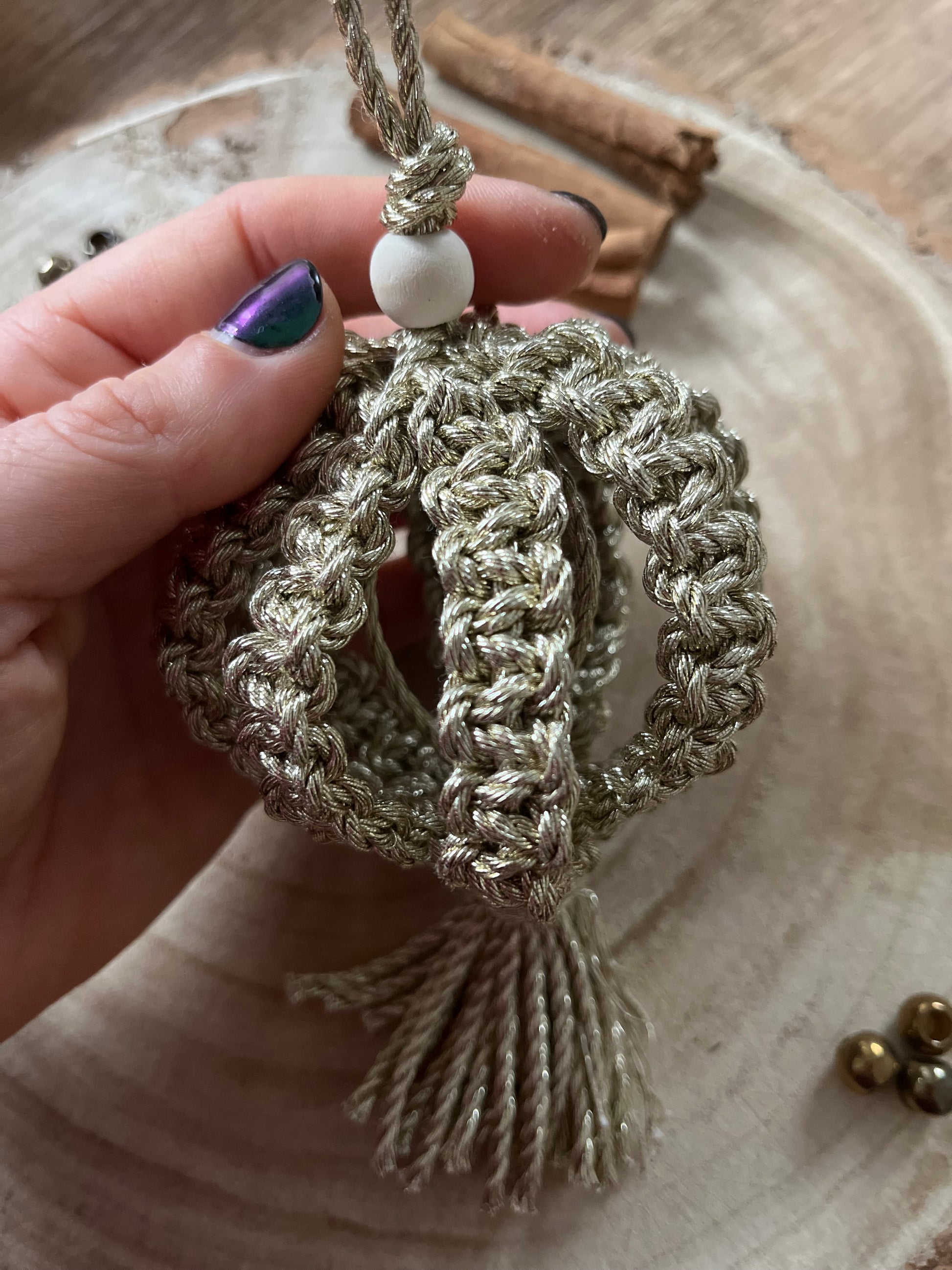 A macrame Christmas bauble made with metallic gold cotton yarn, and finished with a small wooden bead, is held in a person's hand, over a round wooden surface. .