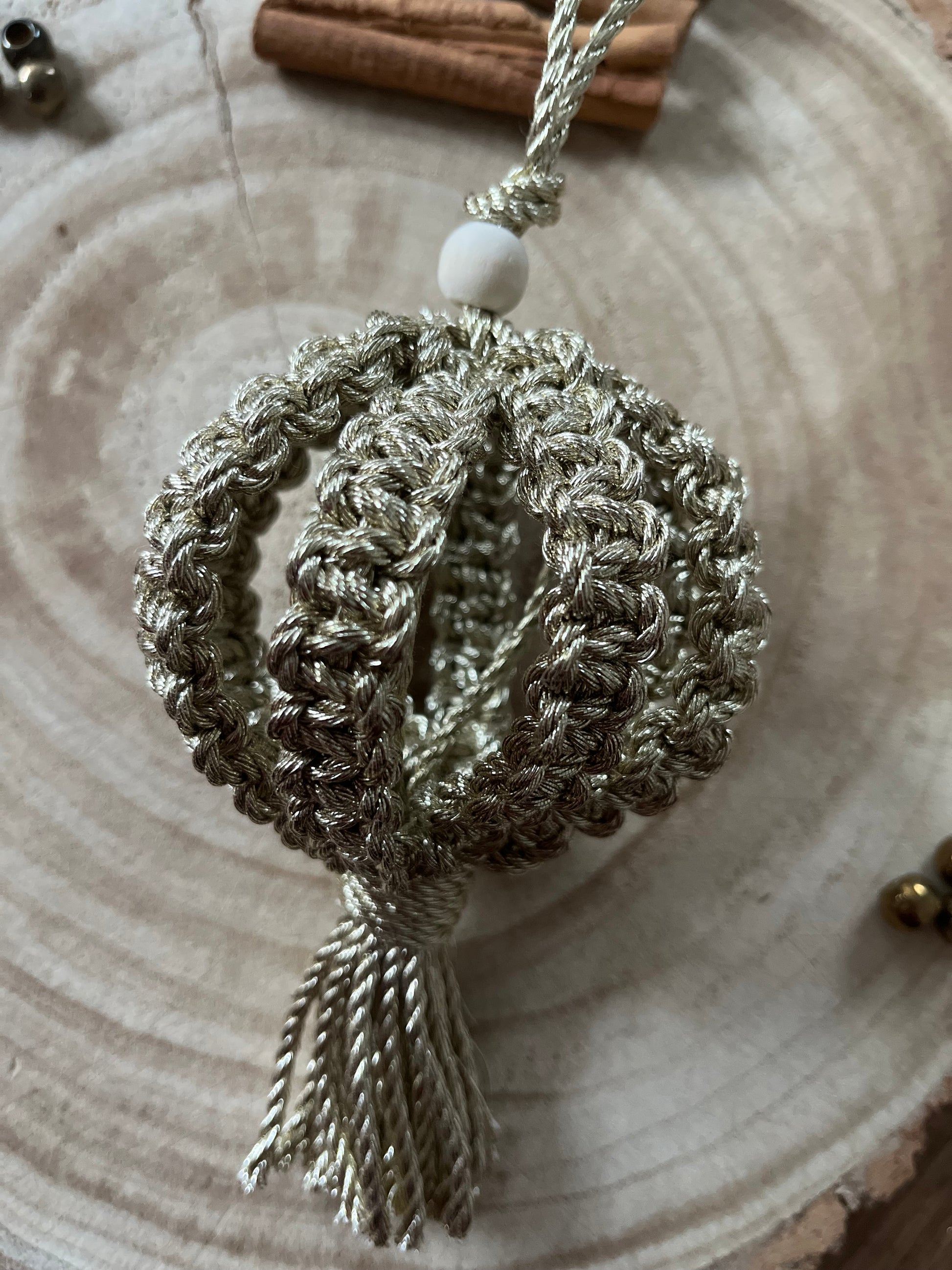 A macrame Christmas bauble made with metallic gold cotton yarn, and finished with a small wooden bead, is placed on a round wooden surface. .