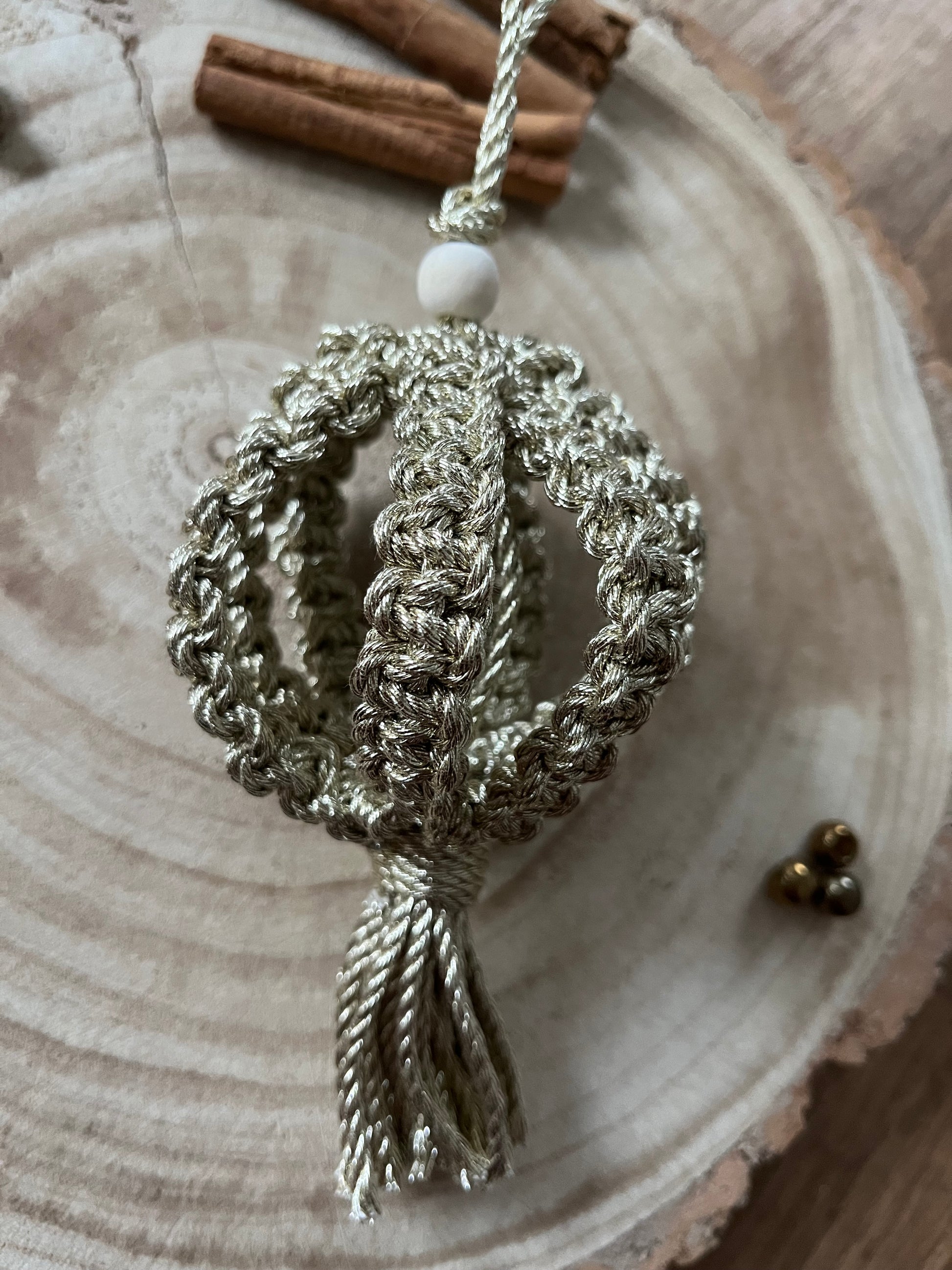 A macrame Christmas bauble made with metallic gold cotton yarn, and finished with a small wooden bead, is placed on a round wooden surface, surrounded by small gold beads and cinnamon sticks.
