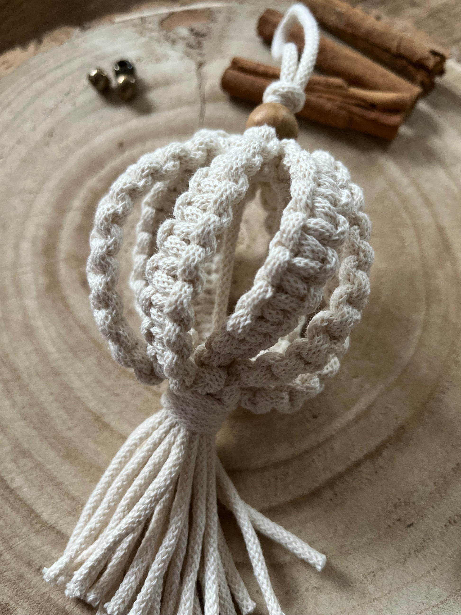 A macrame Christmas bauble made with natural cotton yarn, and finished with a small wooden bead, is placed on a round wooden surface, surrounded by small beads and cinnamon sticks for decoration.