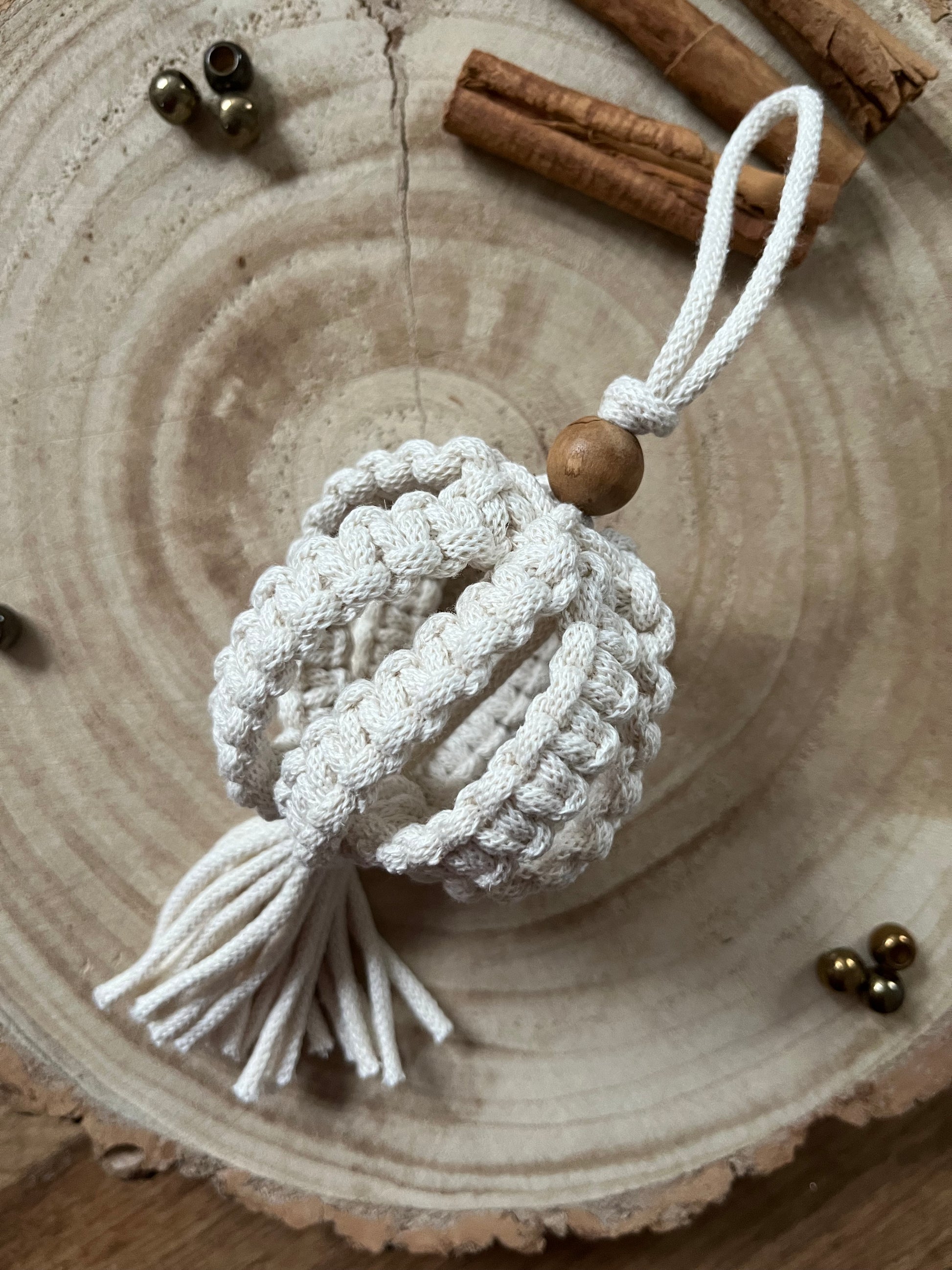 A macrame Christmas bauble made with natural cotton yarn, and finished with a small wooden bead, is placed on a round wooden surface, surrounded by small beads and cinnamon sticks for decoration.