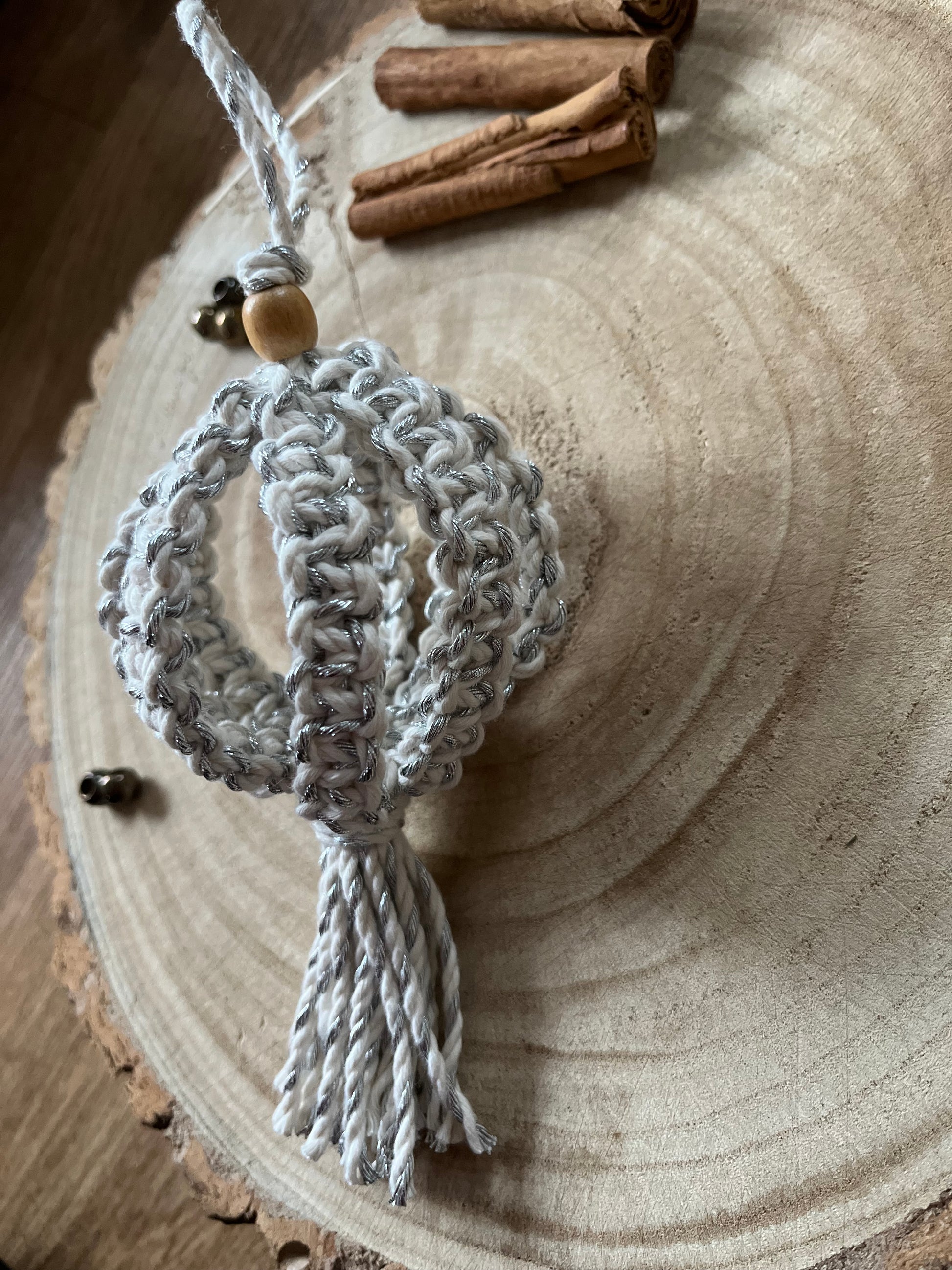 A macrame Christmas bauble made with silver cotton yarn, and finished with a small wooden bead, is placed on a round wooden surface, surrounded by small beads and cinnamon sticks for decoration.