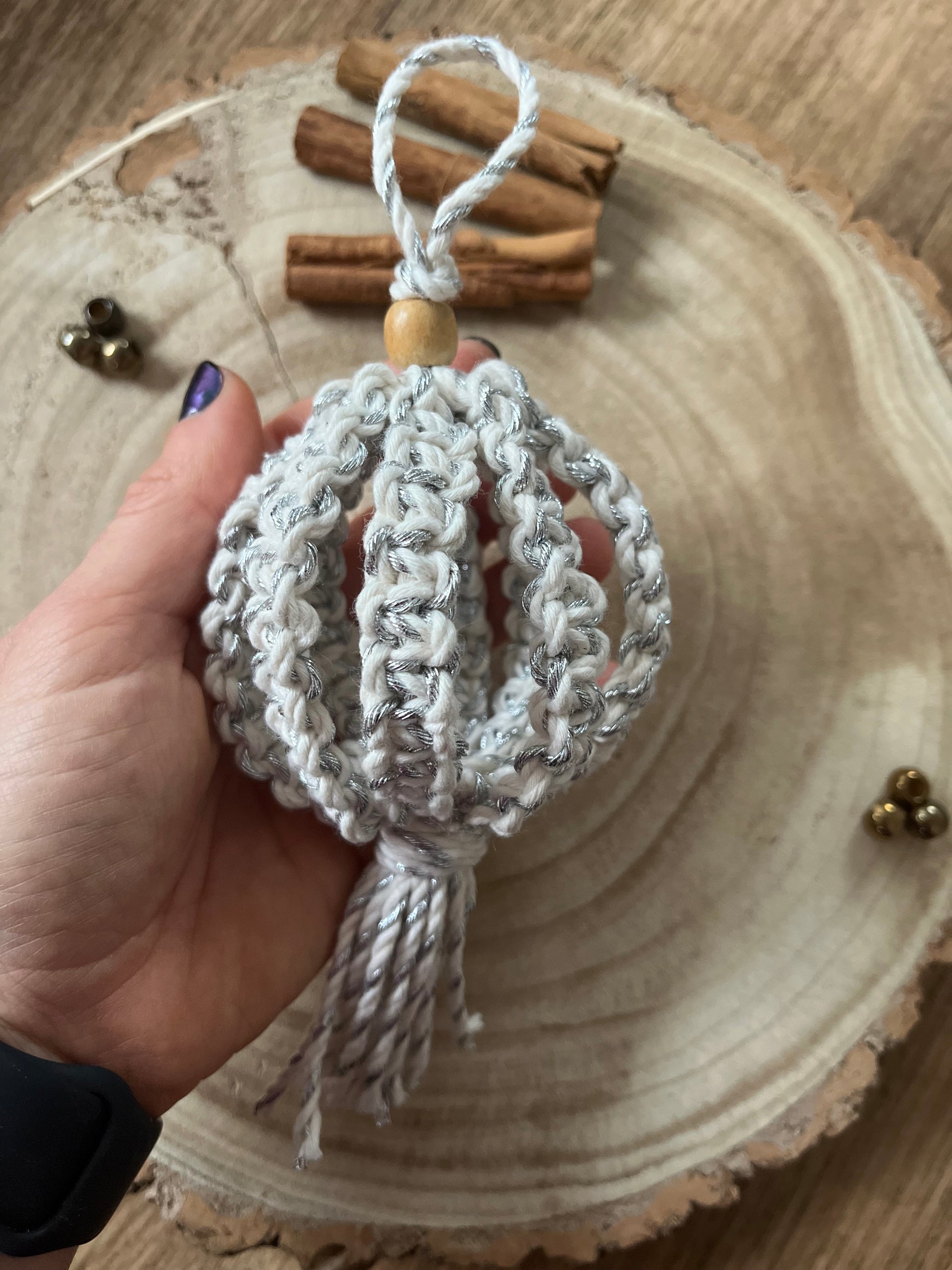 A hand holds a macrame Christmas bauble made with silver cotton yarn, and finished with a small wooden bead.