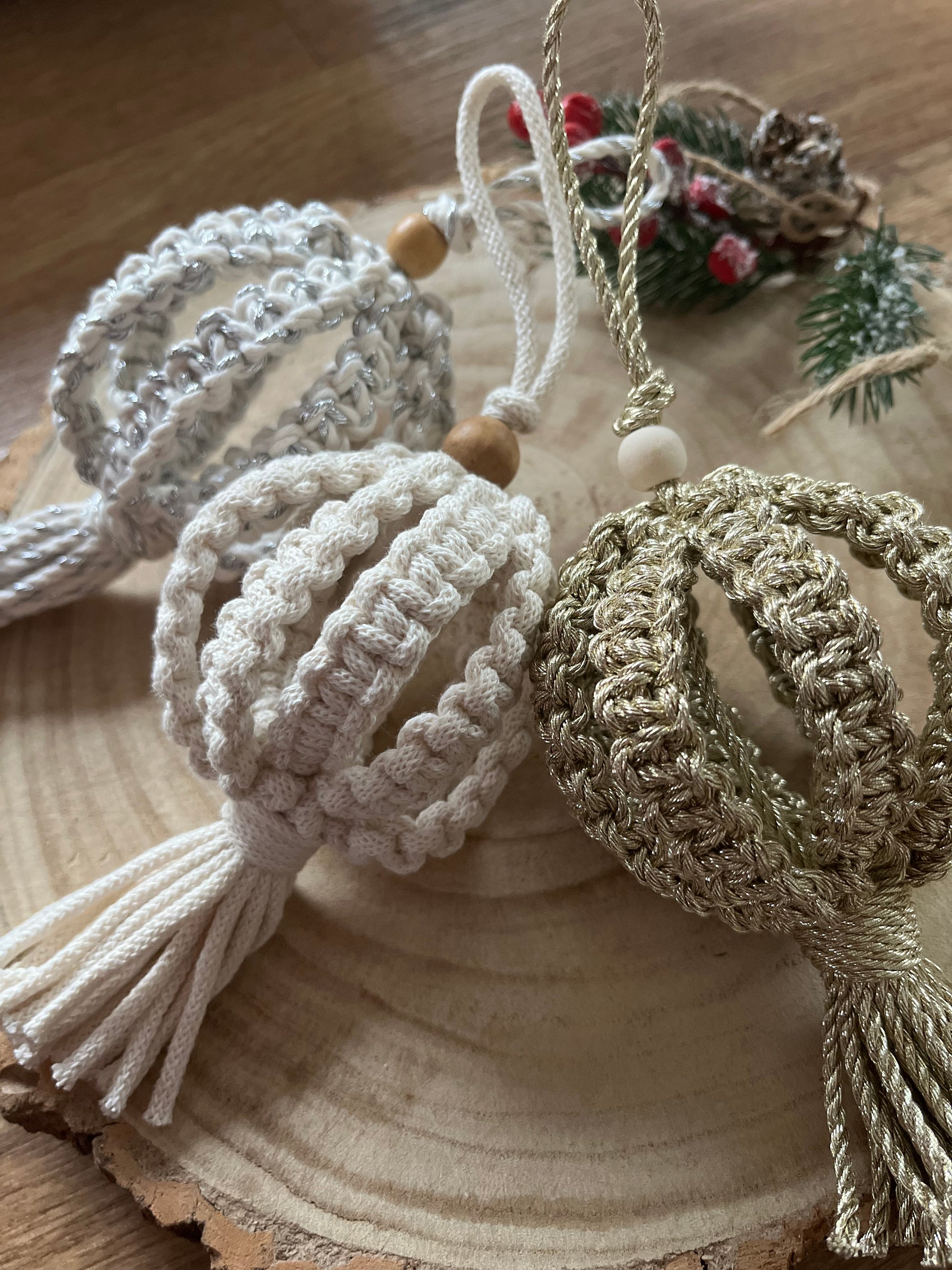 Three knotted Christmas baubles made with silver, natural and gold cotton yarns, and finished with a small wooden bead, lay beside each other on a round wooden surface.