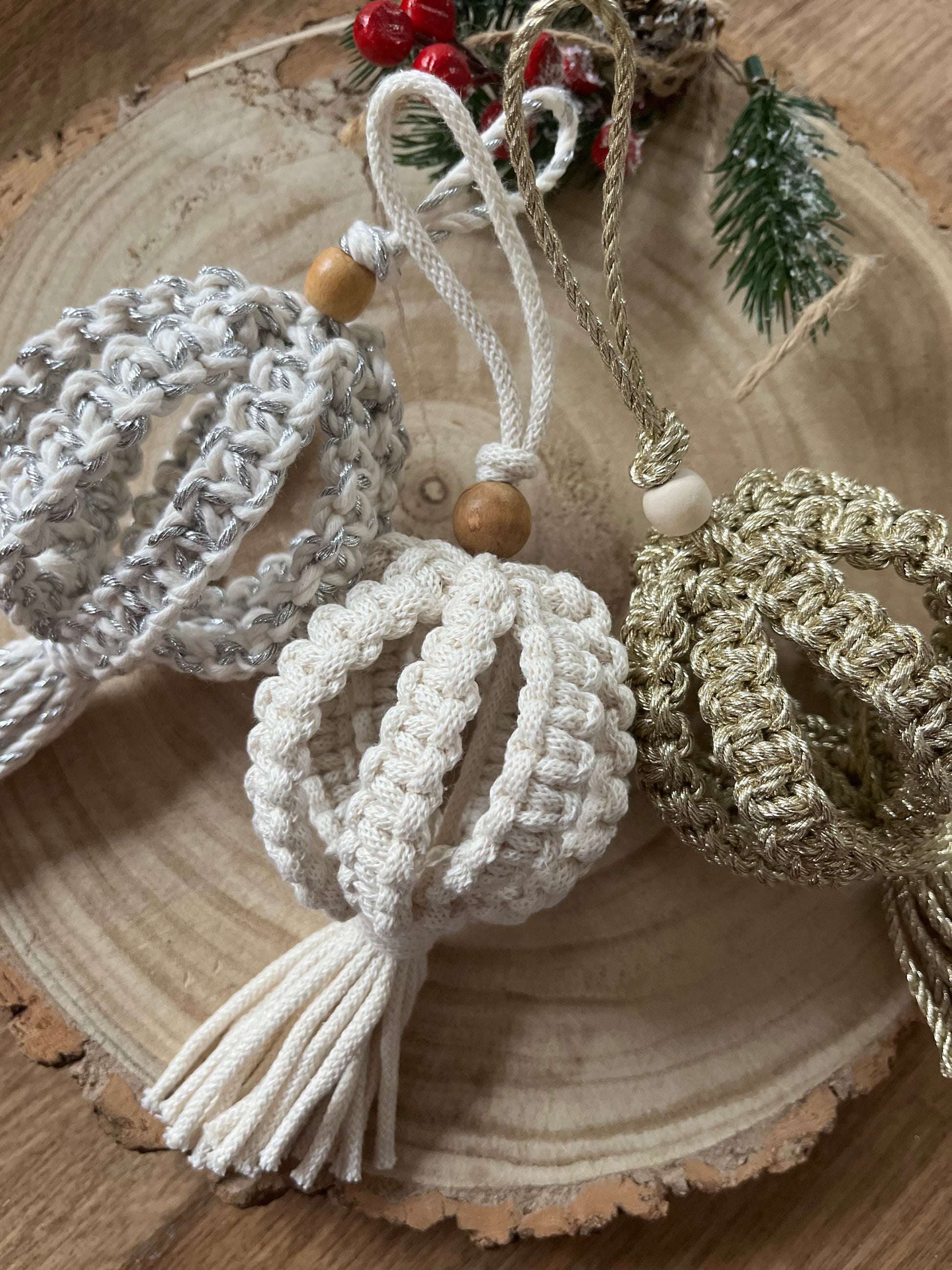 Three knotted Christmas baubles made with silver, natural and gold cotton yarns, and finished with a small wooden bead, lay beside each other on a round wooden surface.