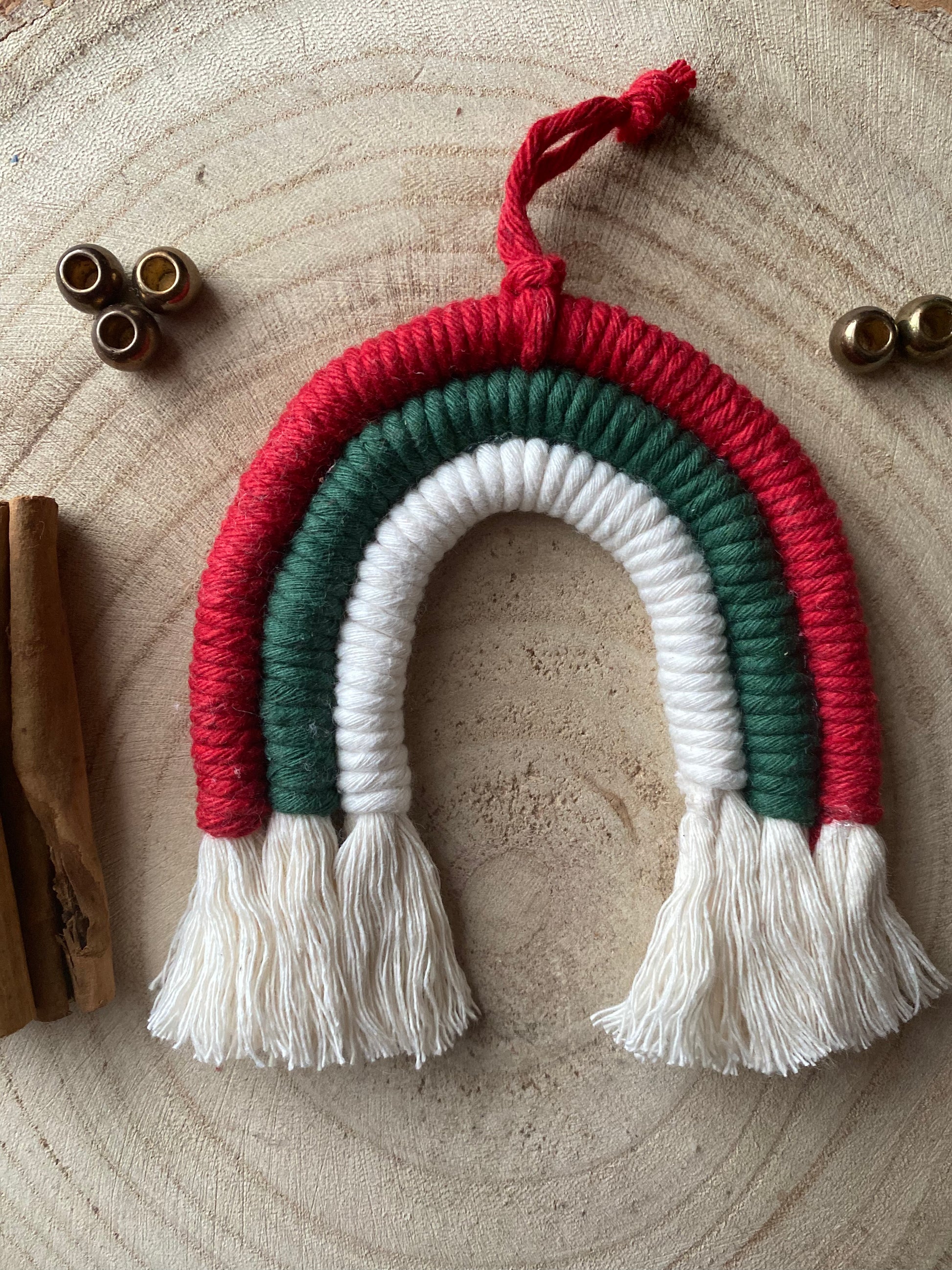 A handmade macrame rainbow Christmas decoration in red, green, and white yarn with white tassels, displayed on a wooden surface with beads and cinnamon sticks for decoration.