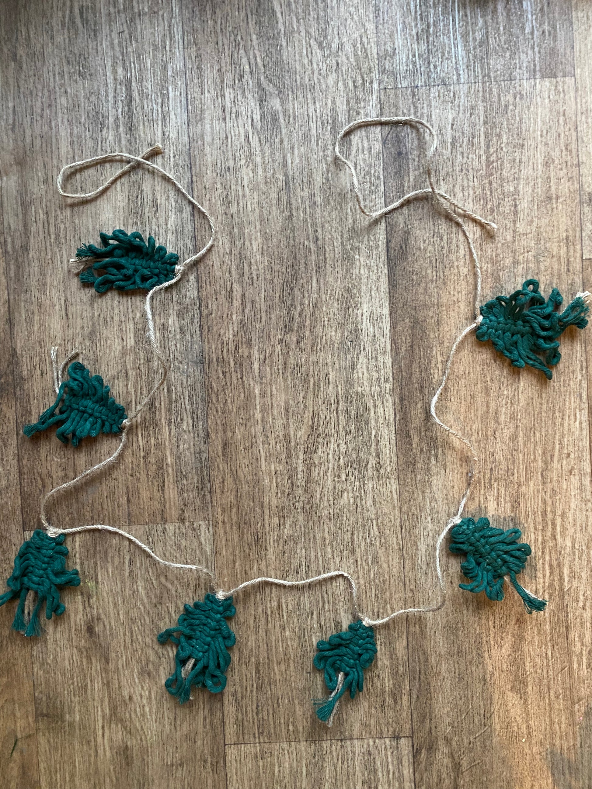 A garland of handmade macrame christmas trees, made with pine green cotton yarn on a length of jute string, laid out on a wooden floor.