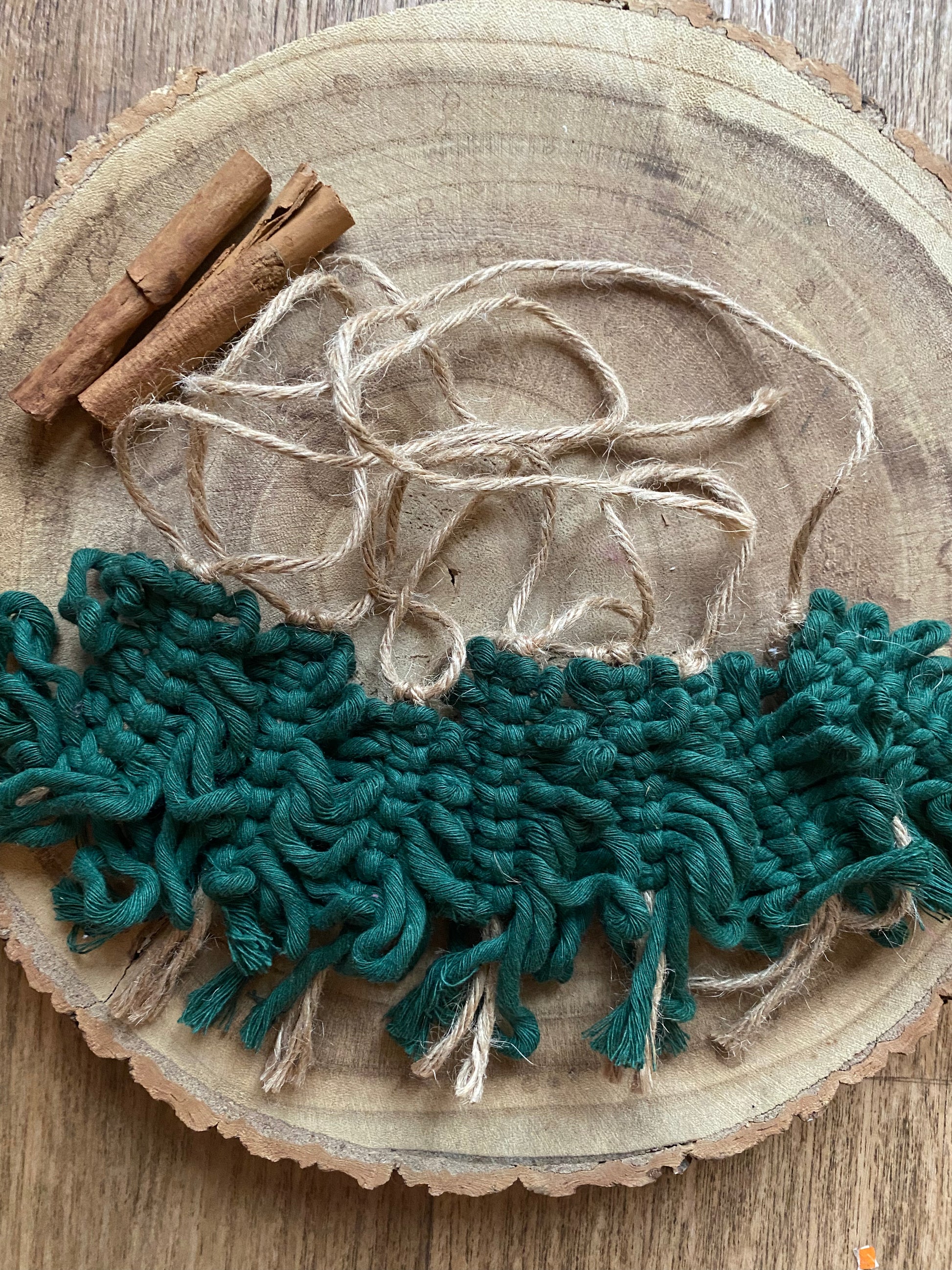 A garland of handmade macrame christmas trees, made with pine green cotton yarn on a length of jute string, displayed neatly on a wooden board, with some cinnamon sticks.