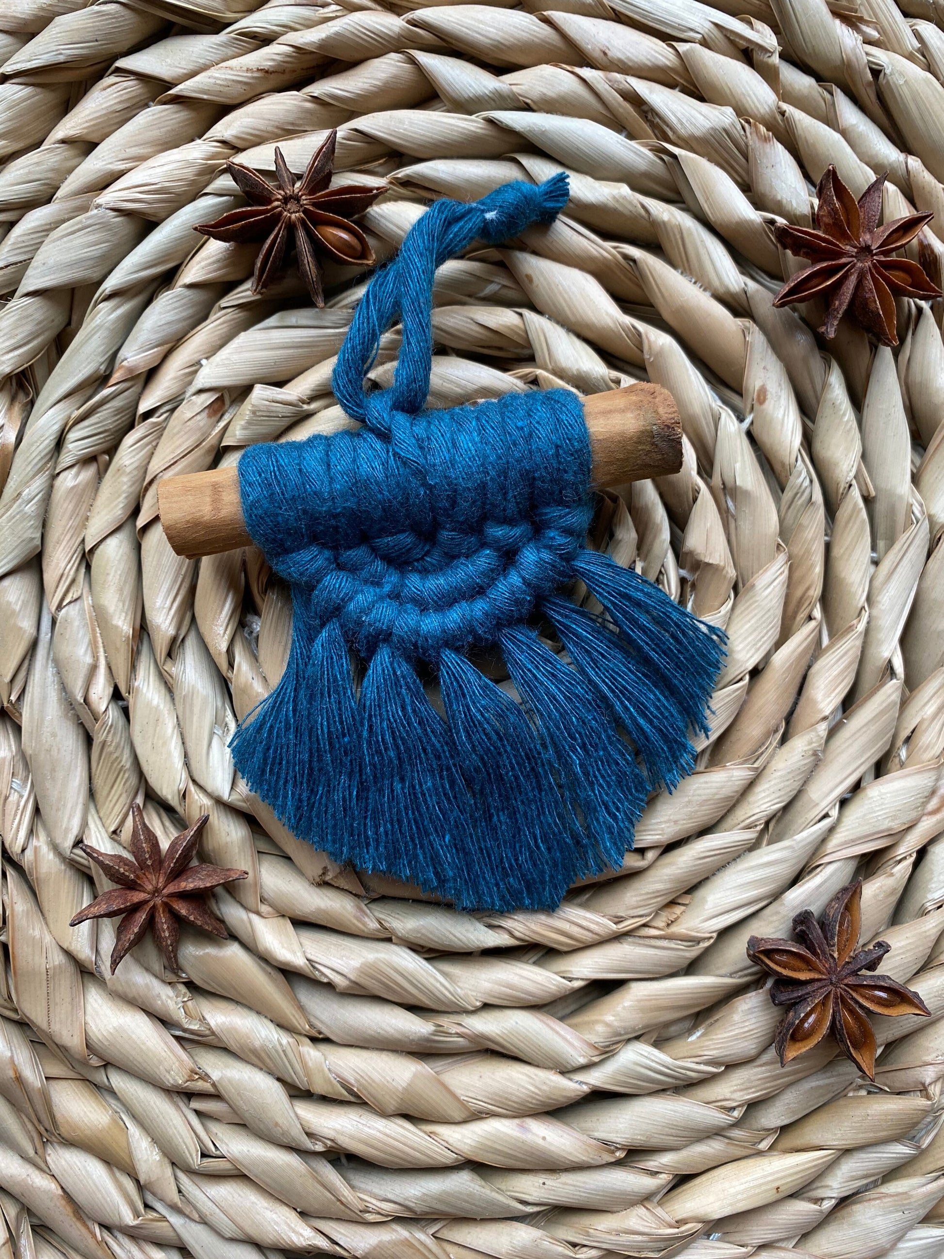 A handmade macrame christmas tree decoration made with blue recycled cotton and a real cinnamon stick, laid out on a round wicker mat surrounded by star anise for decoration.