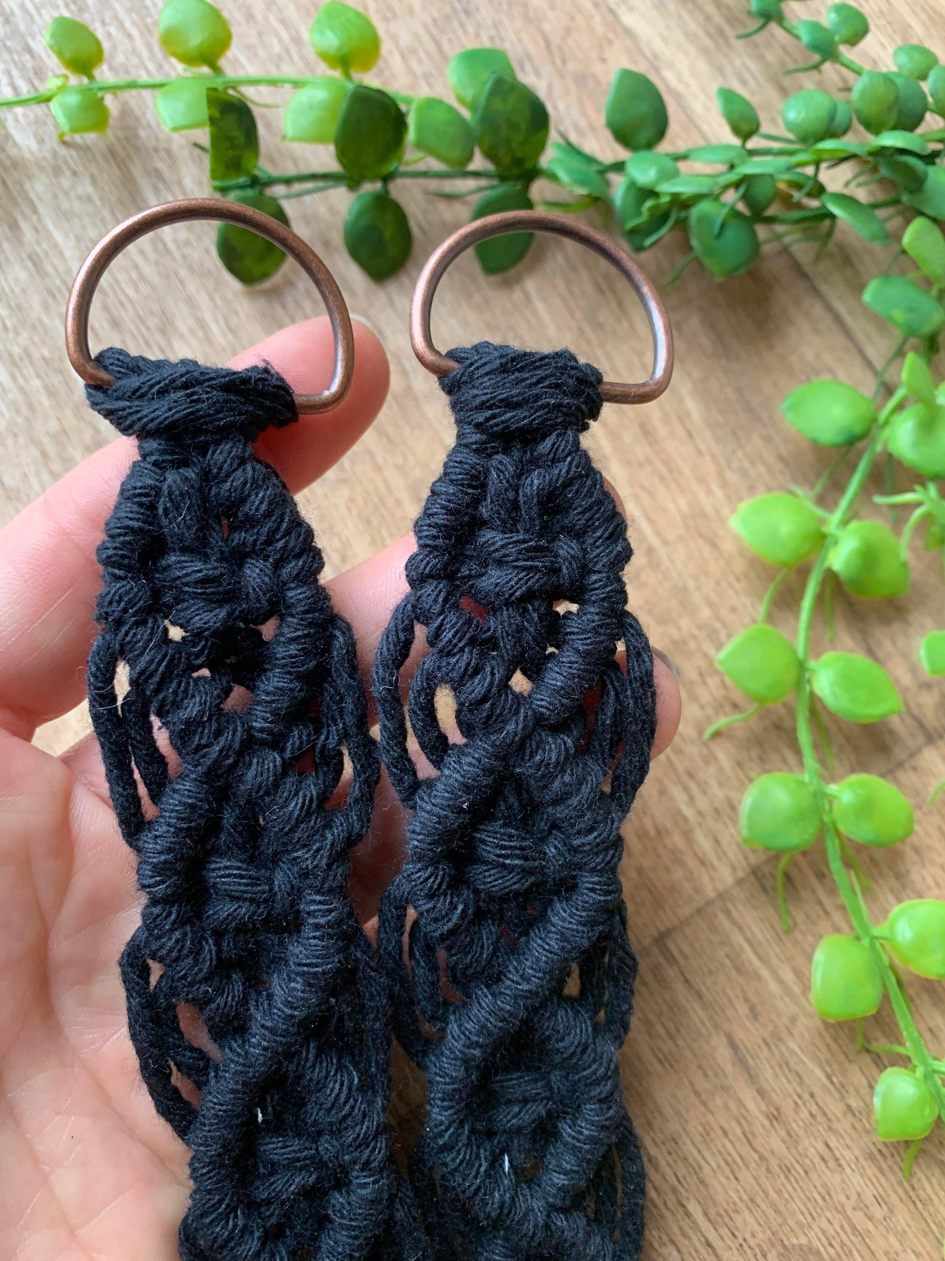 Close-up of a person's hand holding a pair of knotted recycled cotton curtain tie backs featuring intricate knot patterns and metal D rings, made with black yarn.