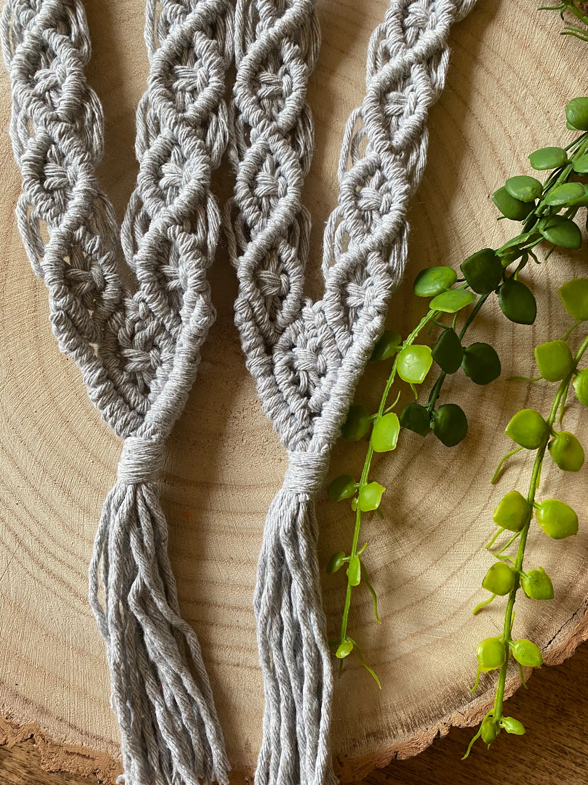 A close-up of a pair of light grey coloured knotted recycled cotton curtain tie backs featuring intricate macramé design with diamond patterns, lying on a wooden surface next to green plant vines.
