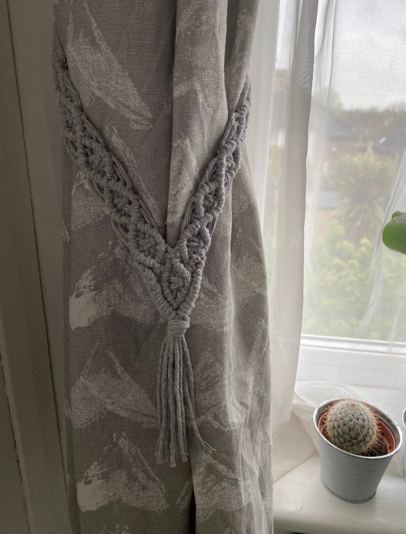 A close-up view of a grey curtain tied back with a pair of knotted recycled cotton curtain tie backs, also in light grey. The curtain hangs beside a window adorned with a white net curtain, and a small potted cactus sits on the windowsill.