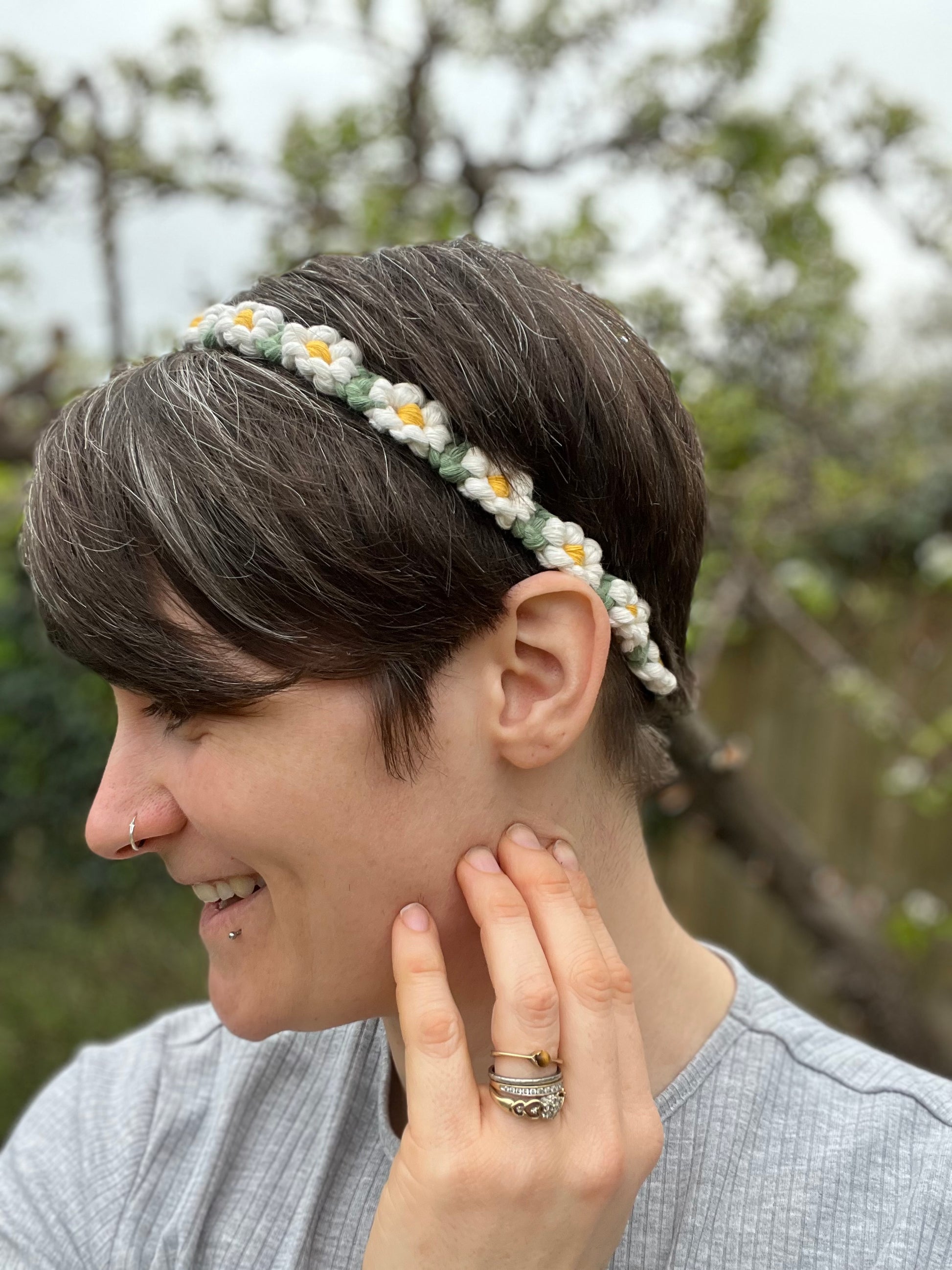 A woman with short brown hair wearing a daisy chain headband with a smile on her face, sideways to the camera, set against an outdoor backdrop.