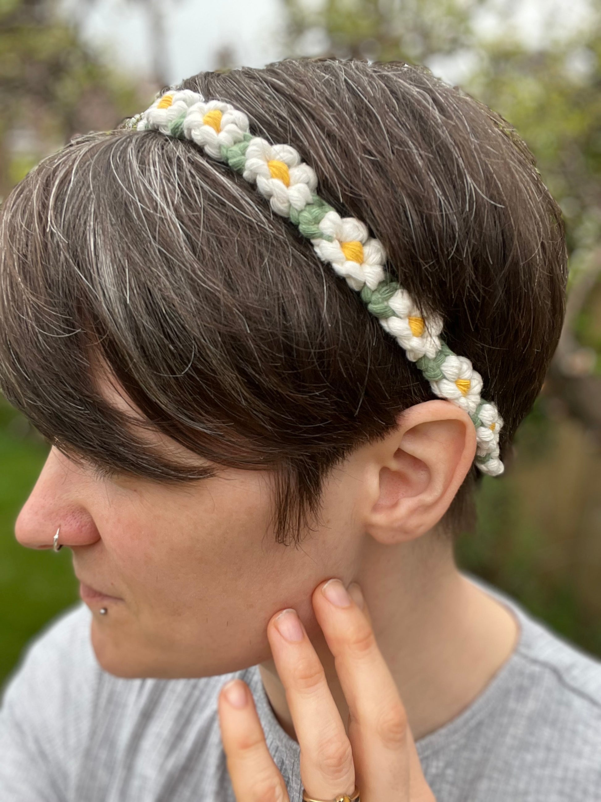 A woman with short brown hair wearing a daisy chain headband sideways to the camera, set against an outdoor backdrop.
