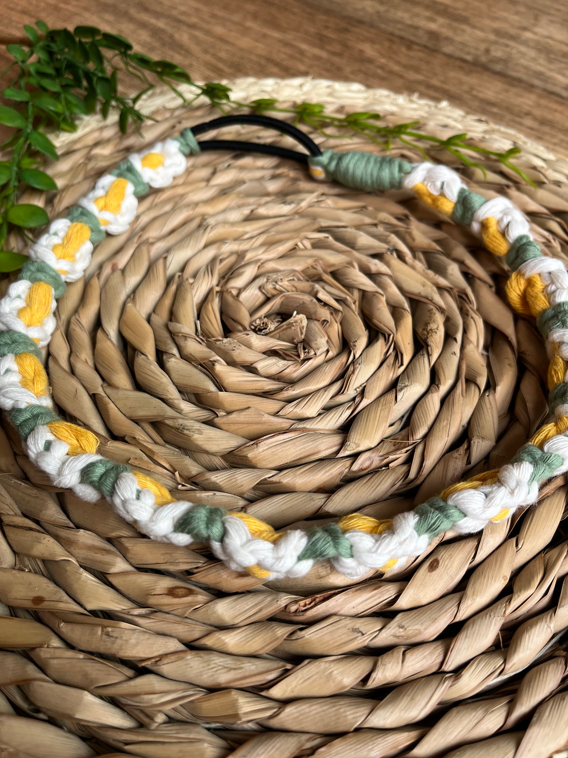 Close up of a macrame daisy chain knotted headband made with white yellow and green cotton, laid neatly on a woven placemat.