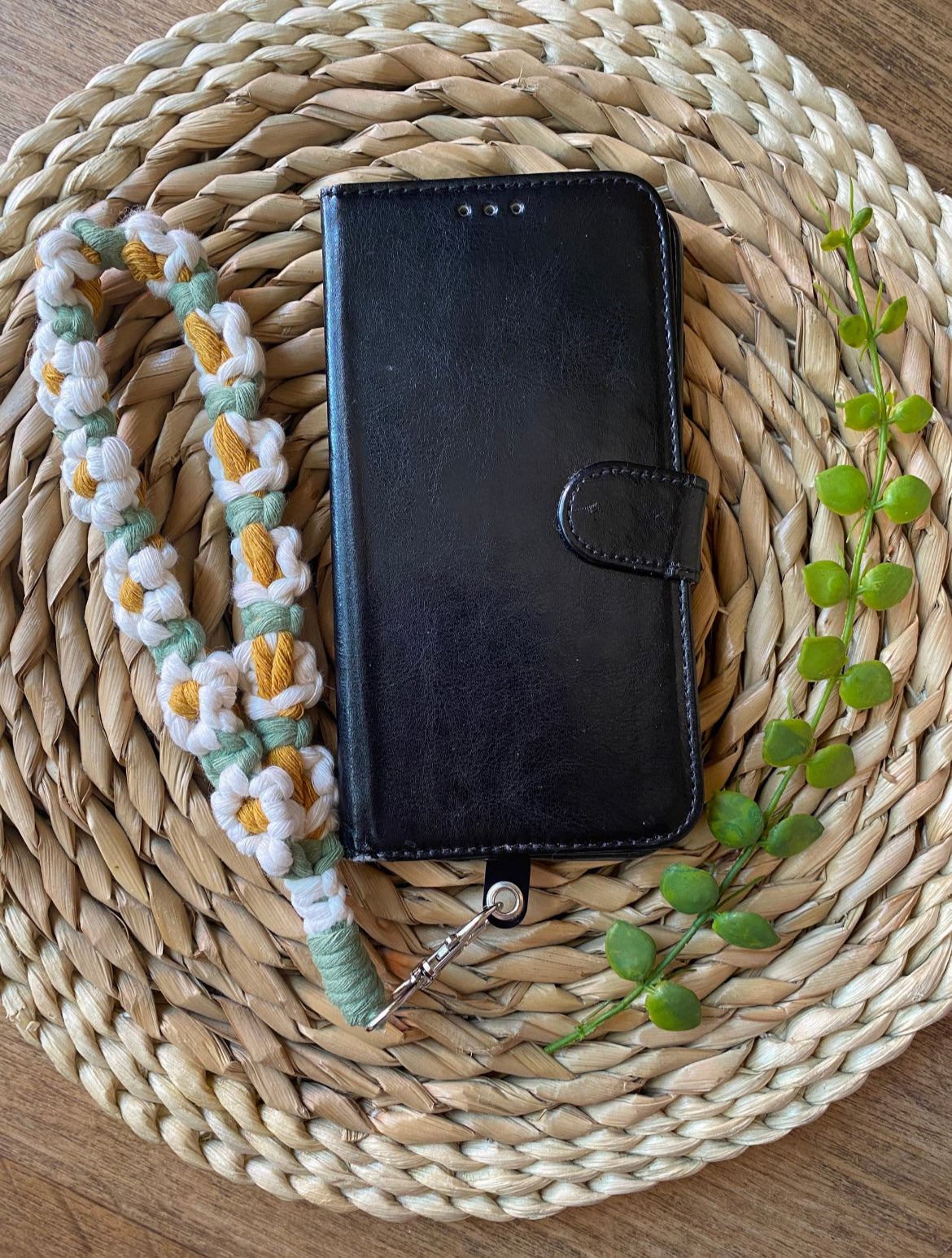 A wristlet phone charm with a daisy chain design laid out next to a black leather phone case on a woven mat.