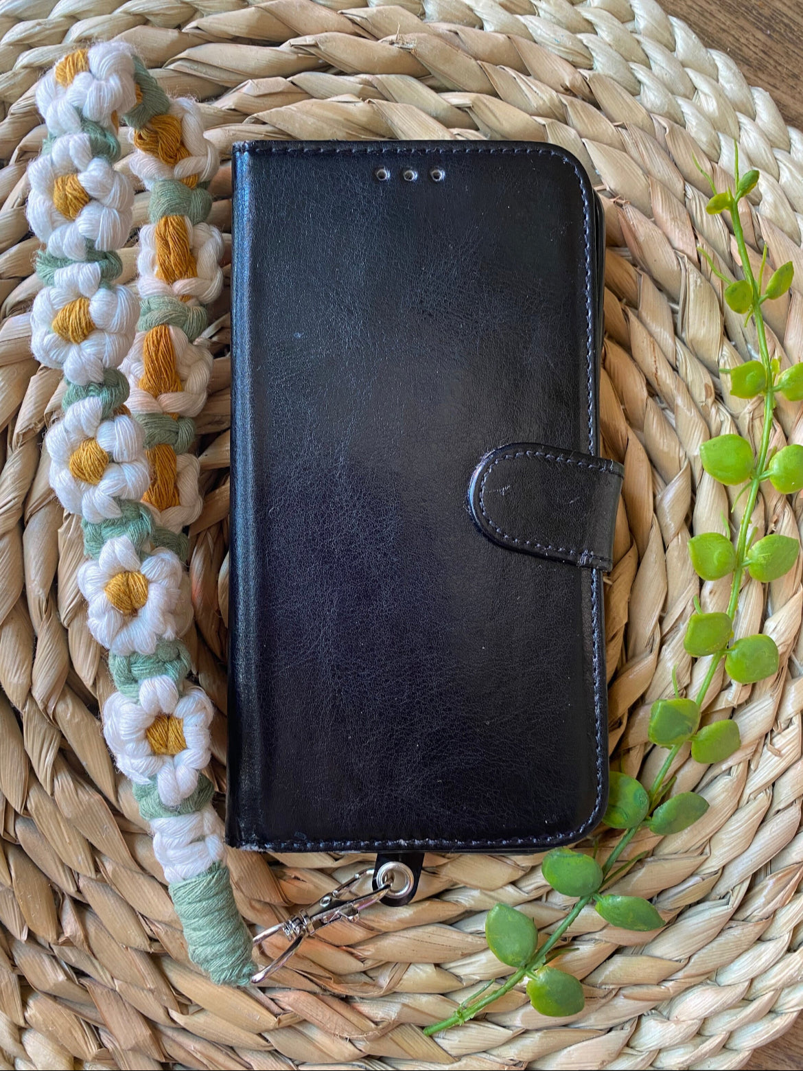 A wristlet phone charm with a daisy chain design laid out next to a black leather phone case on a woven mat.