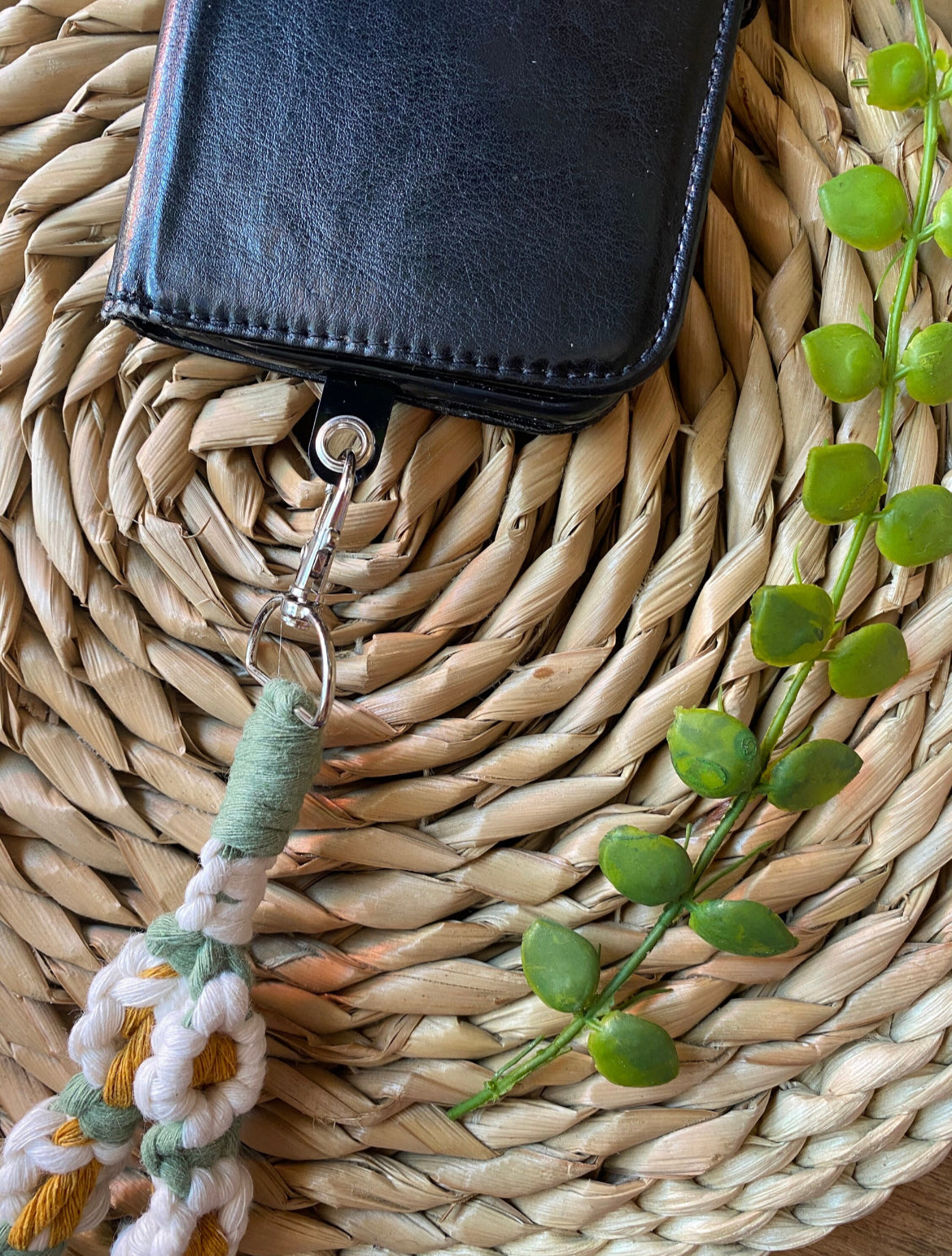 A wristlet phone charm with a daisy chain design attached to a black leather phone case on a woven mat.