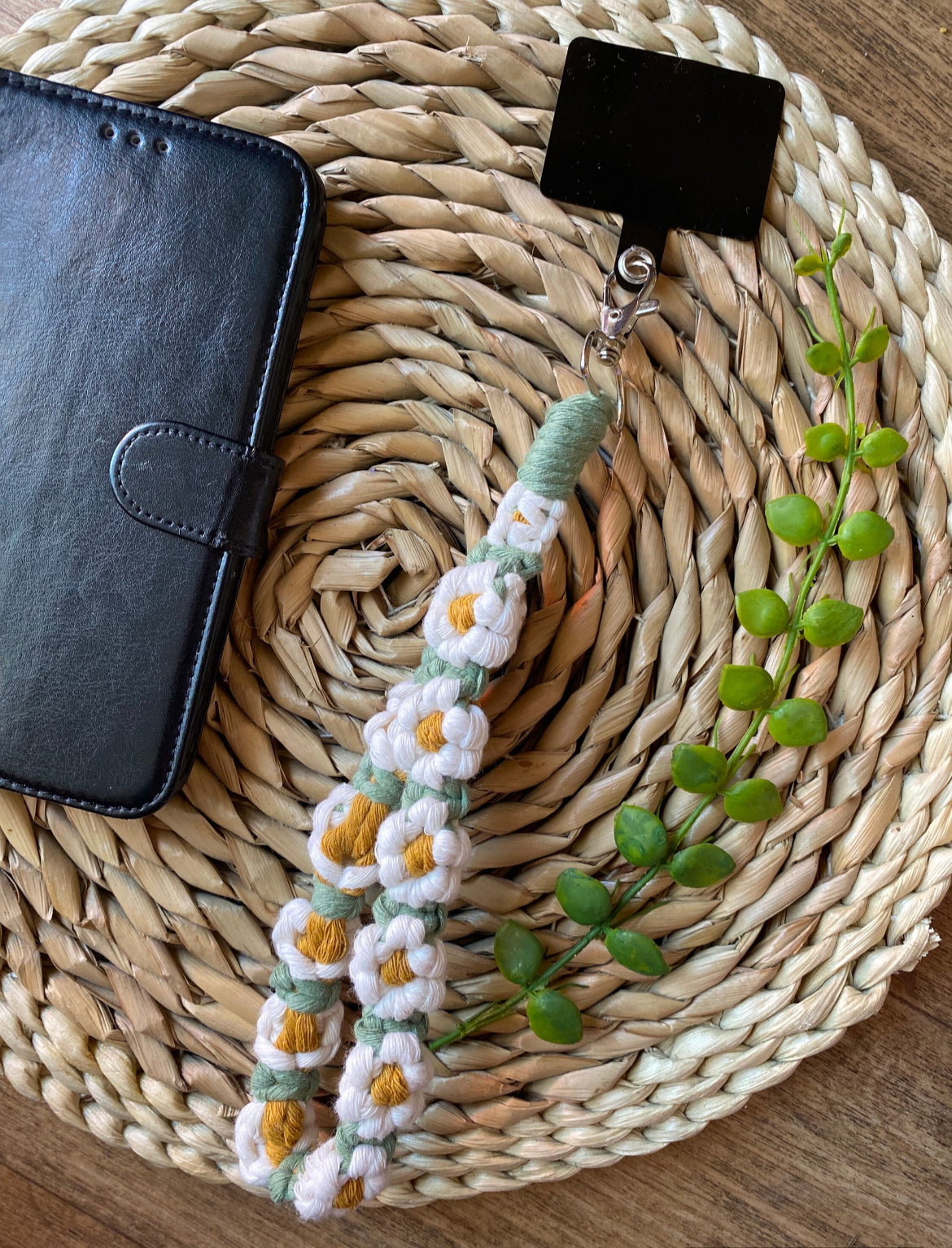 A wristlet phone charm with a daisy chain design, attached to a universal phone tether, laid out next to a black leather phone case on a woven mat.