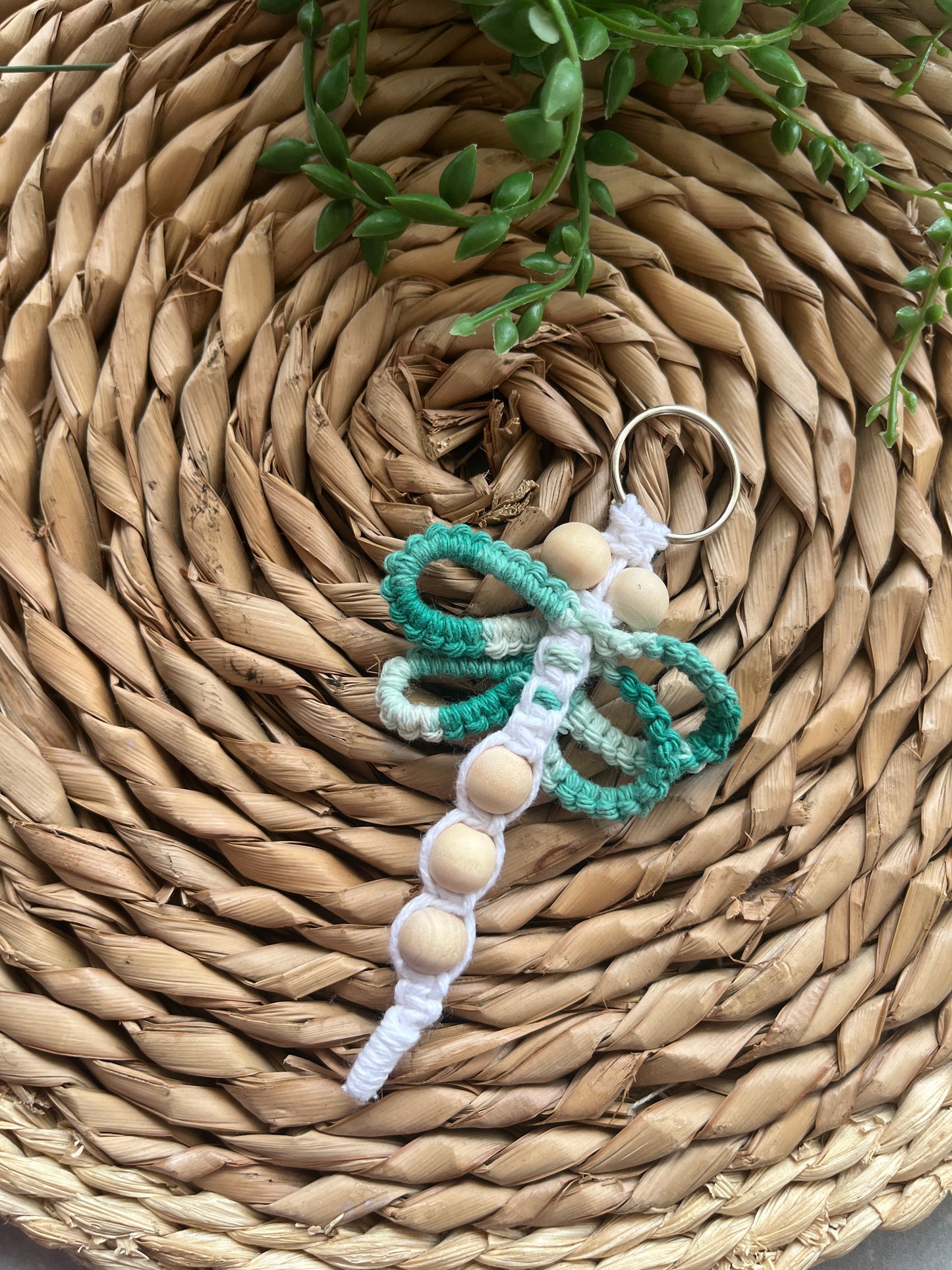 Handmade macrame keychain in the shape of a dragonfly, knotted with mint green and white yarn and small wooden beads, laid on a woven mat.