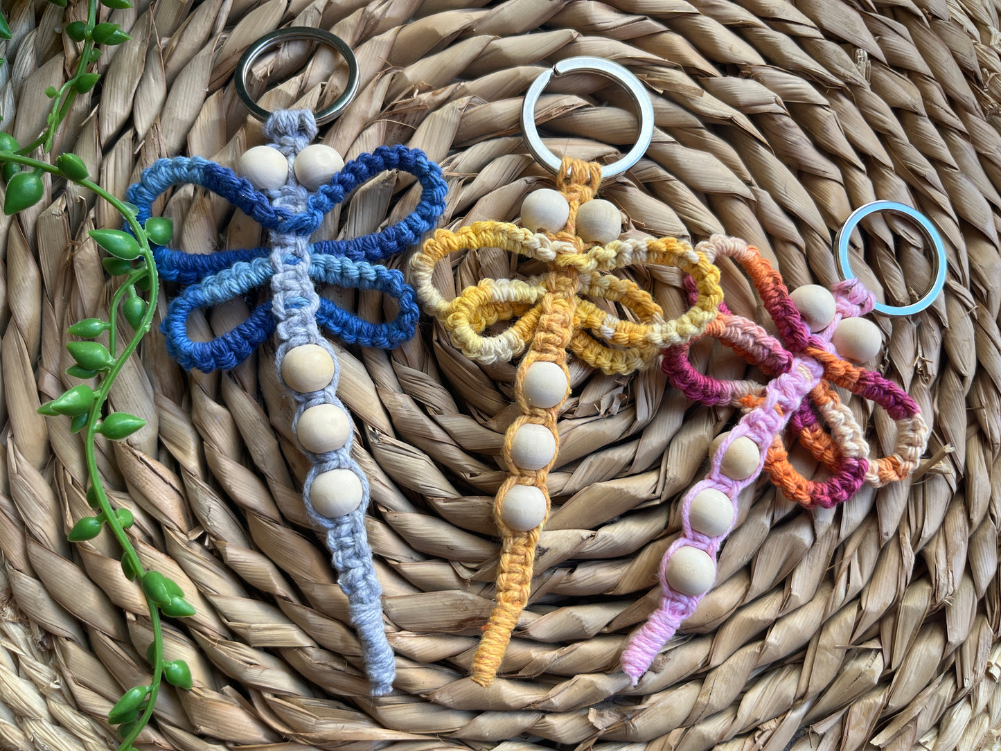 Three handmade macrame dragonfly keyrings, made using shades of blue, yellow and pink recycled cotton, on a metal split ring, with small wooden beads.