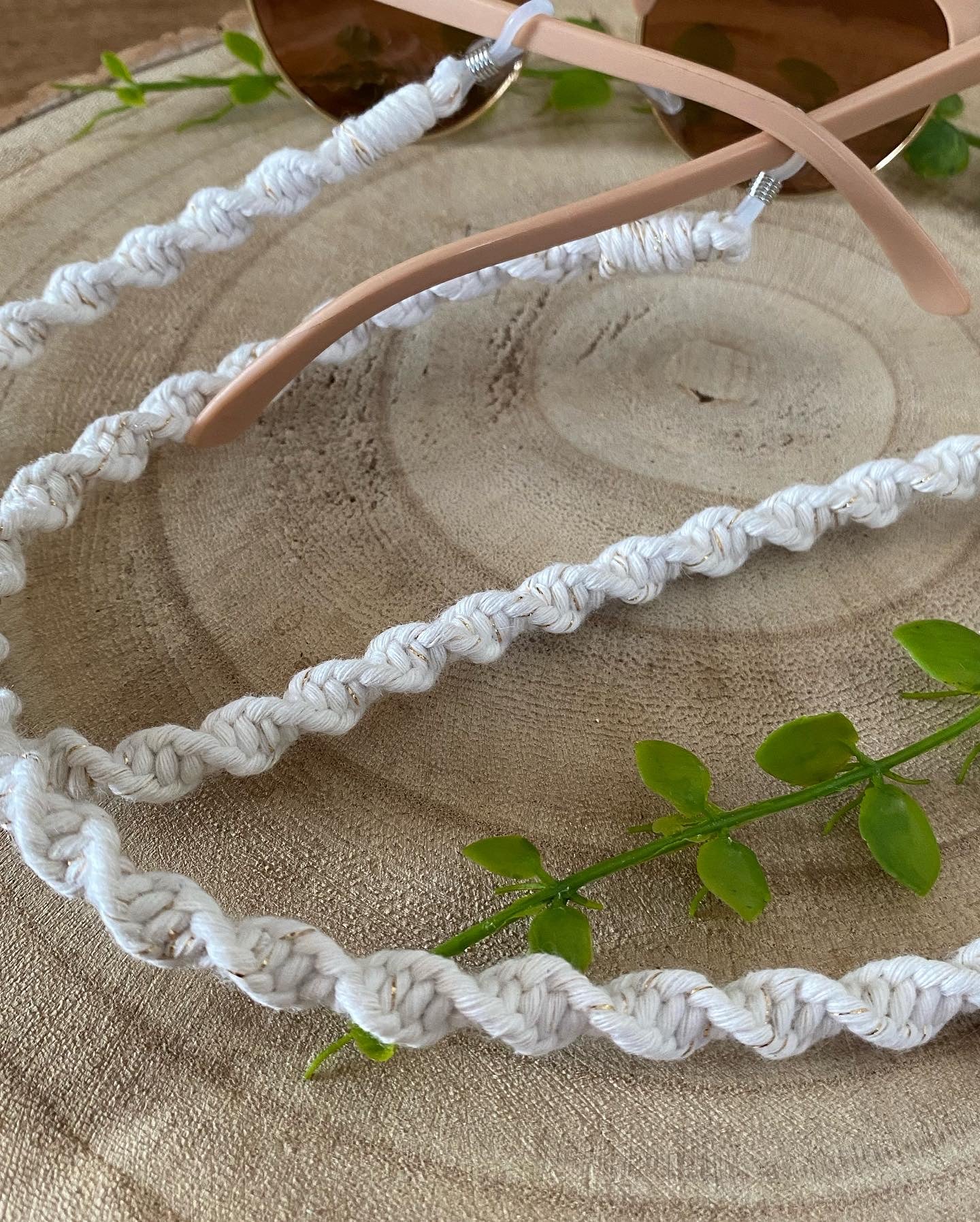 A pair of dark sunglasses are placed on a wooden surface with a handmade macrame eyewear holder chain, made with golden natural recycled cotton yarn, attached to them.