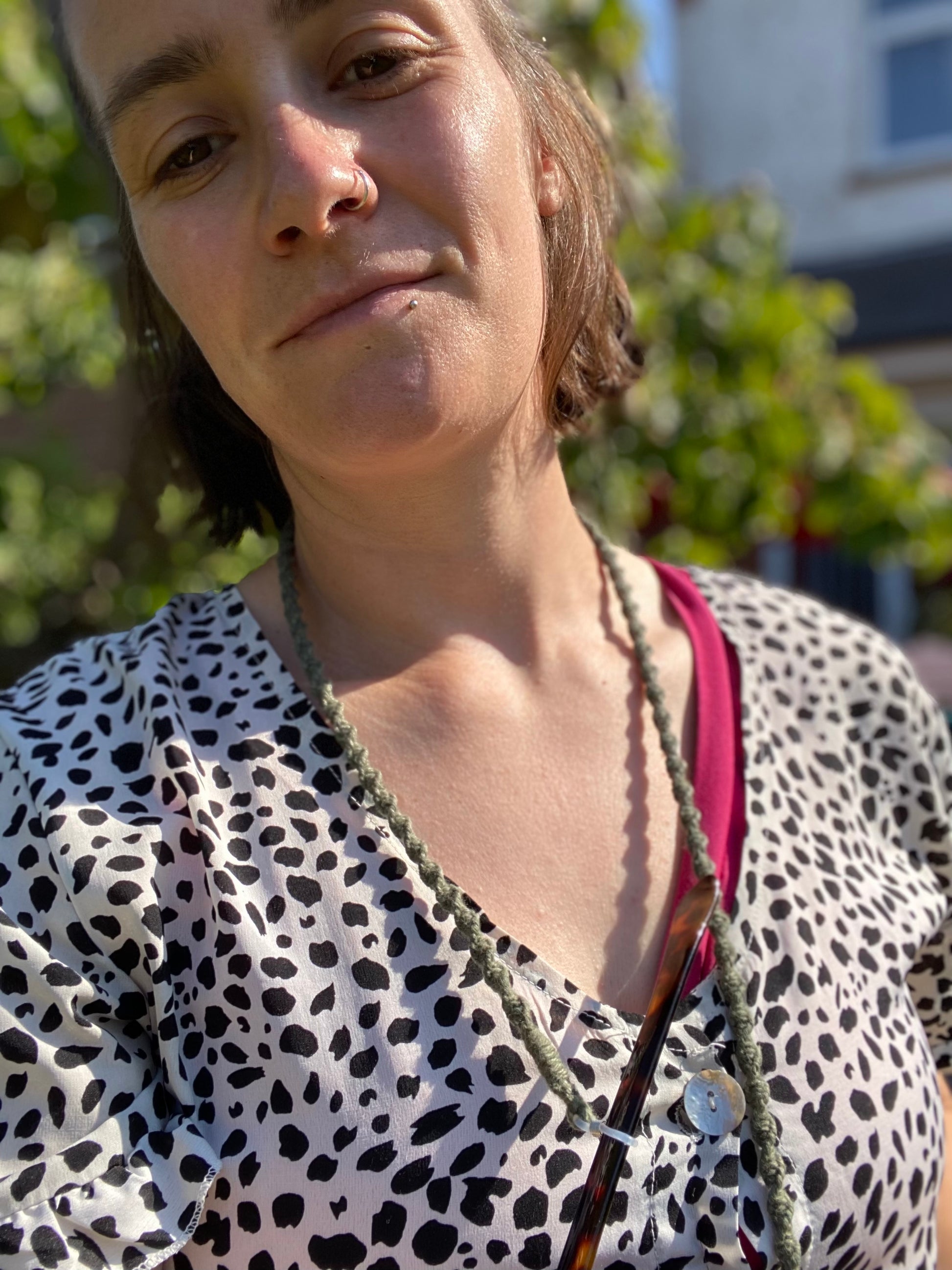 A person with short hair and a nose ring is standing outdoors, wearing a white shirt with black spots and a pink top underneath. They have a relaxed expression and a handmade glasses chain made with olive green recycled cotton around their neck.