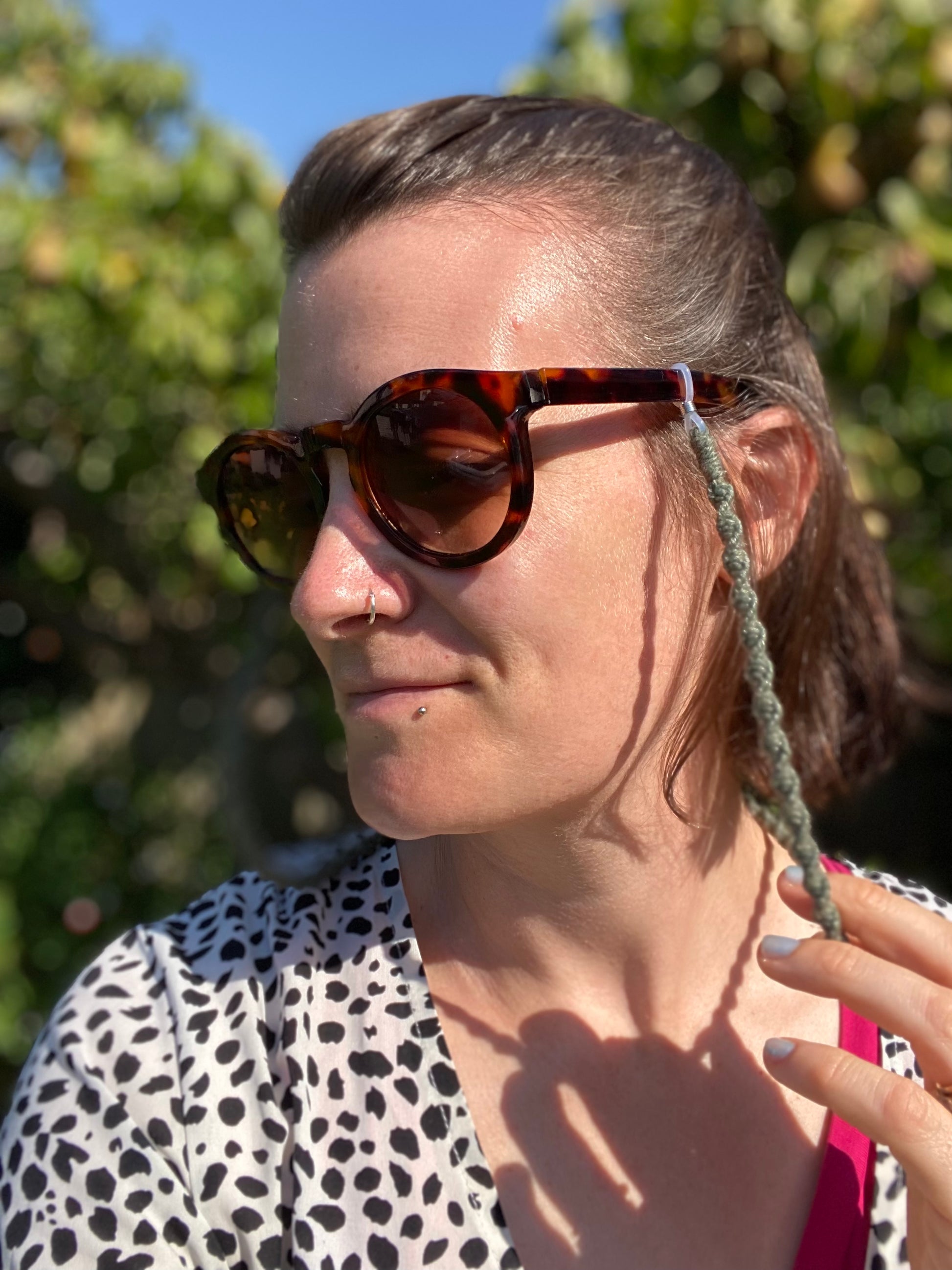 A woman with short hair wearing a pair of dark sunglasses with a handmade macrame eyewear holder chain attached to them stands outside, looking away from the camera.