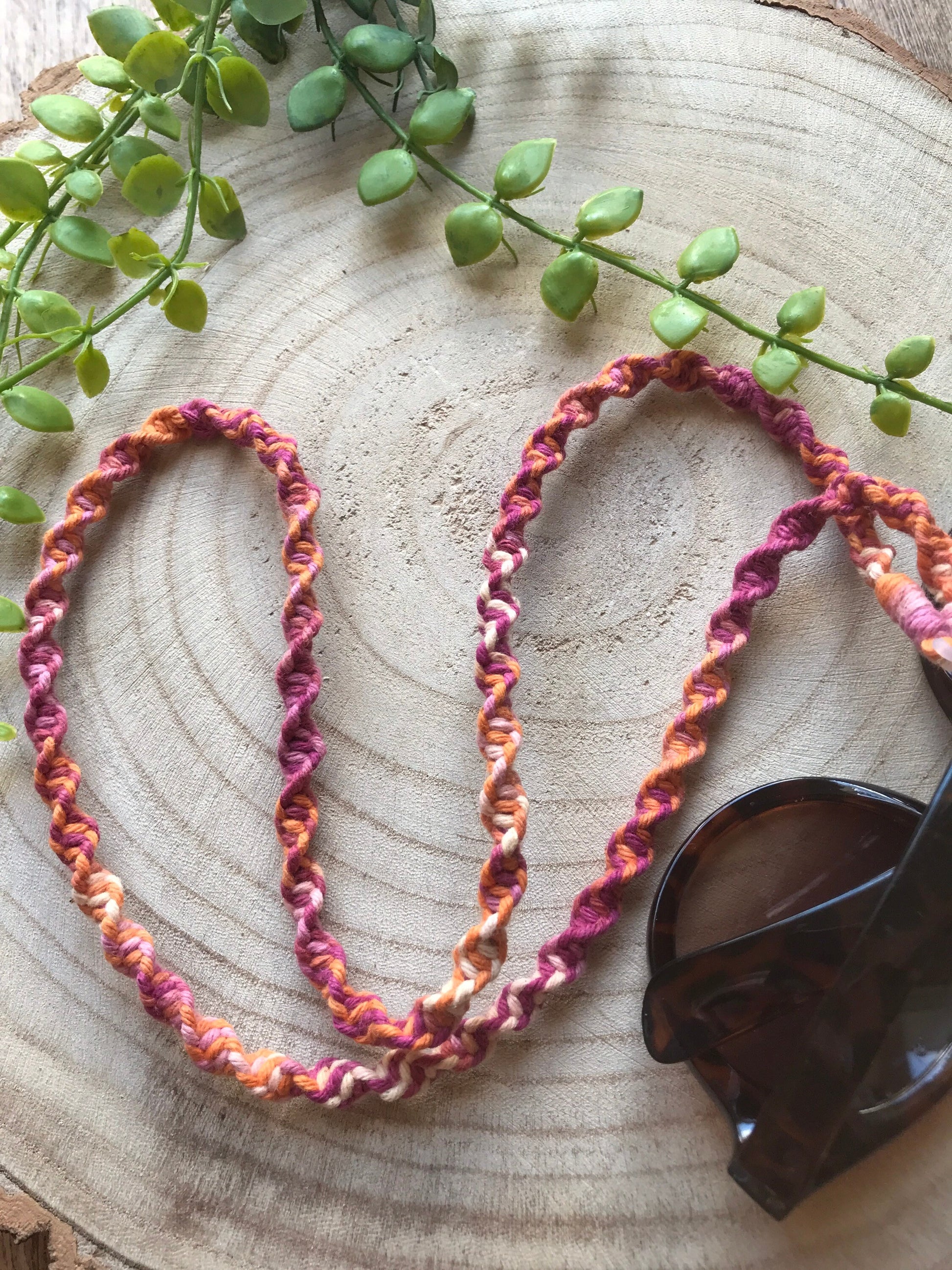 A pair of dark sunglasses are placed on a wooden surface with a handmade macrame eyewear holder chain, made with variegated recycled cotton yarn, attached to them.