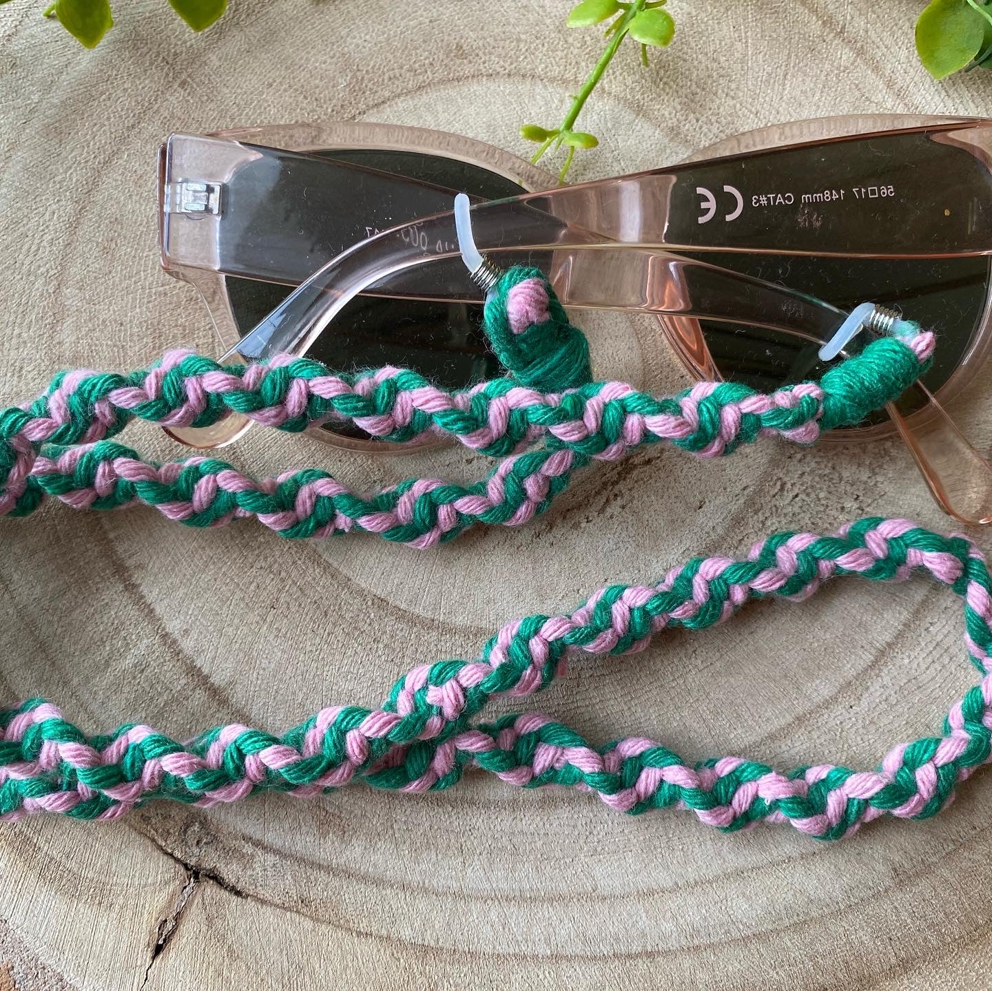 A pair of dark sunglasses are placed on a wooden surface with a handmade macrame eyewear holder chain, made with pink and green recycled cotton yarn, attached to them.